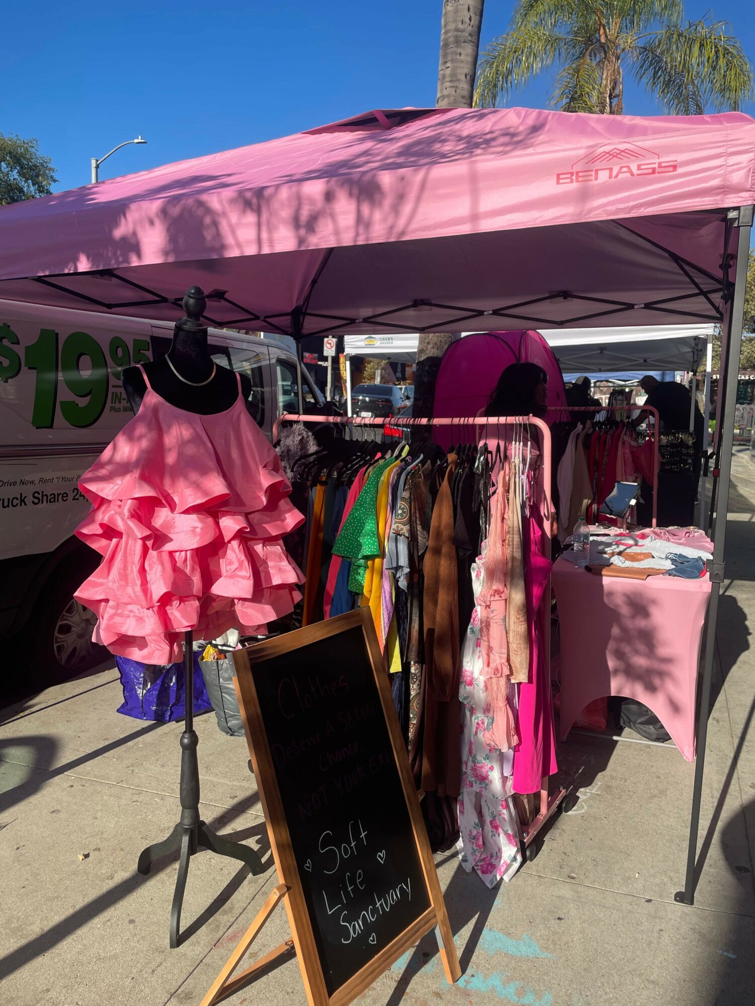 Clothing rack with colorful garments and a pink ruffled dress under a pink canopy, outdoor setting with a clear blue sky.