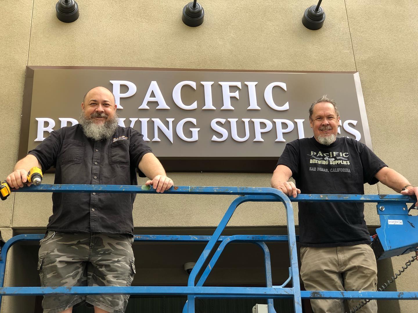 Two men stand on a blue lift platform in front of a sign that reads 'PACIFIC BREWING SUPPLIES.'