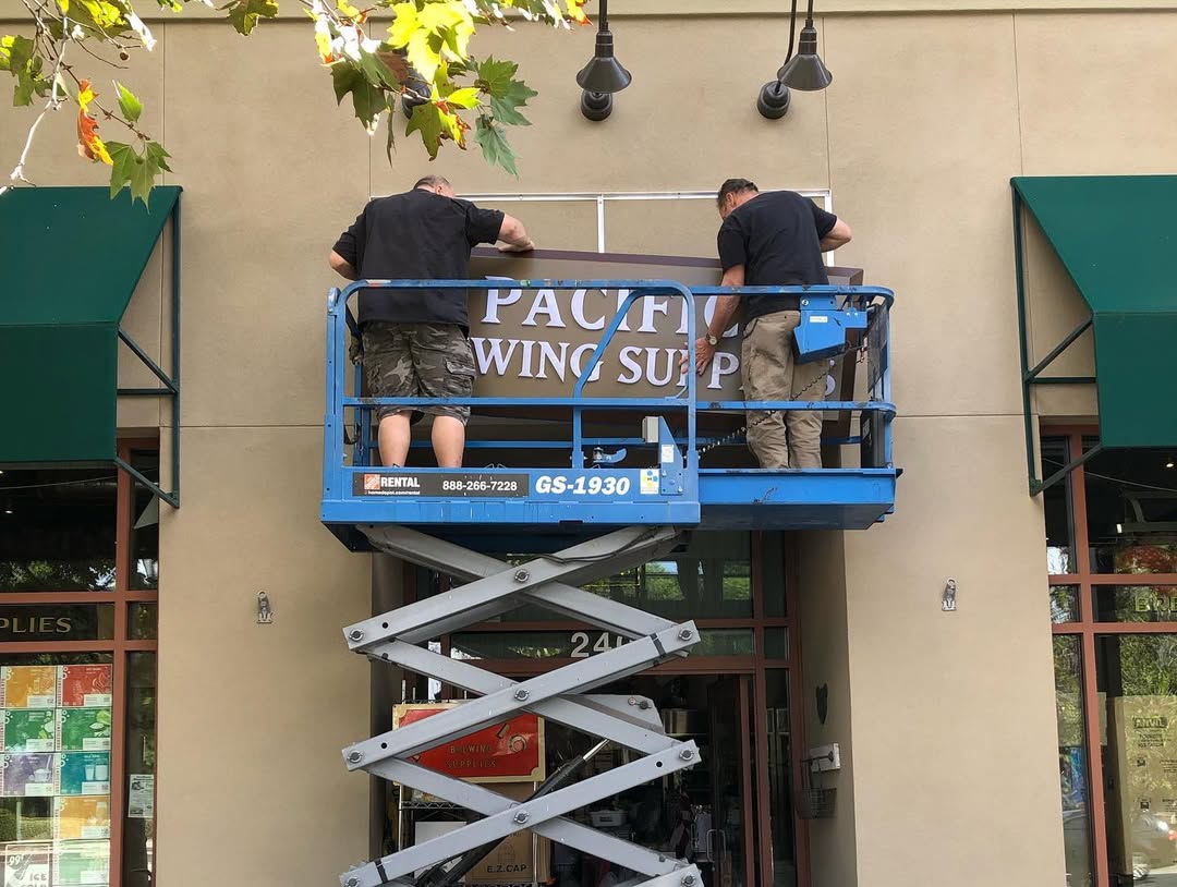 Two workers on a blue lift working on a sign above a storefront entrance.
