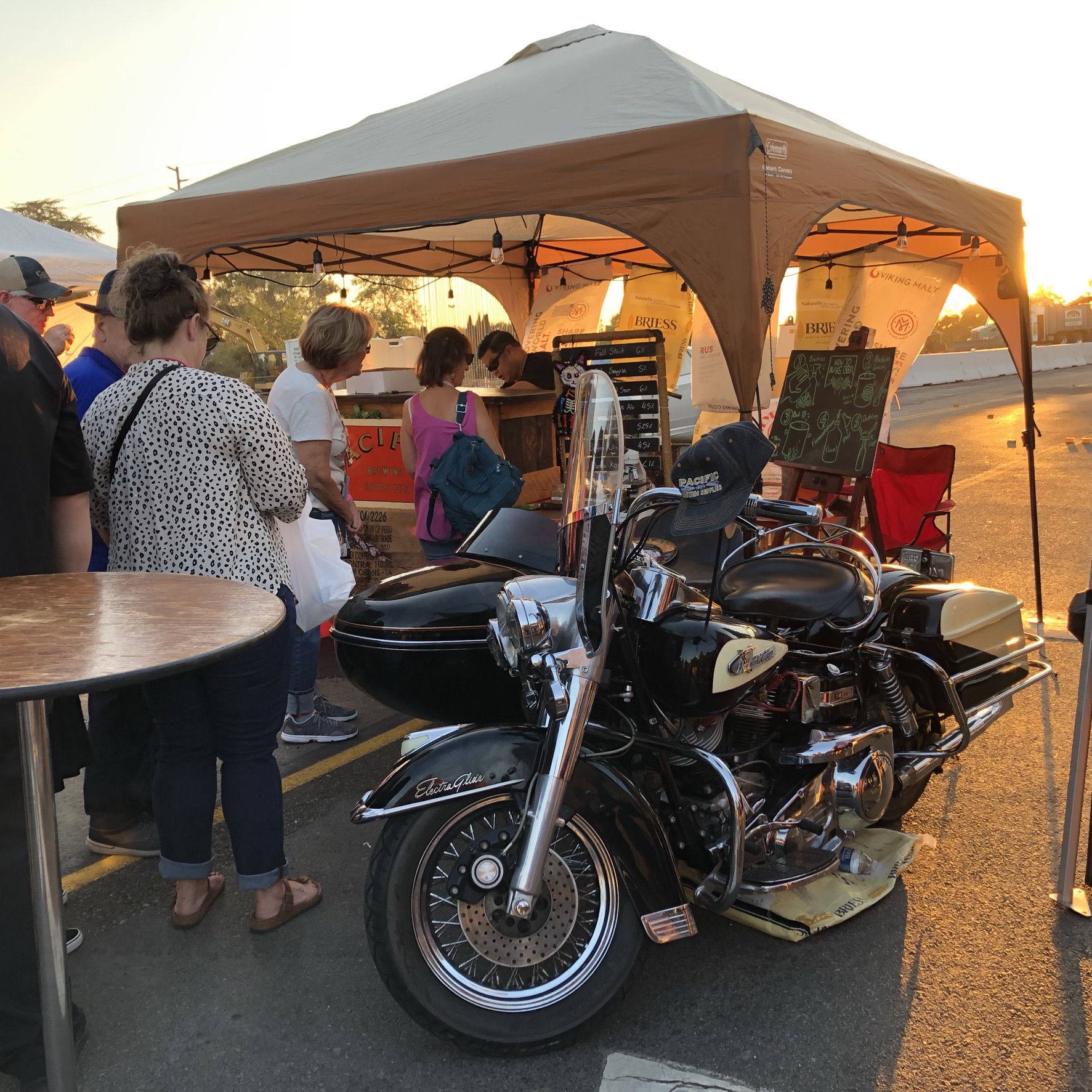Group of people gathered around a booth under a canopy, with a motorcycle parked nearby during sunset.