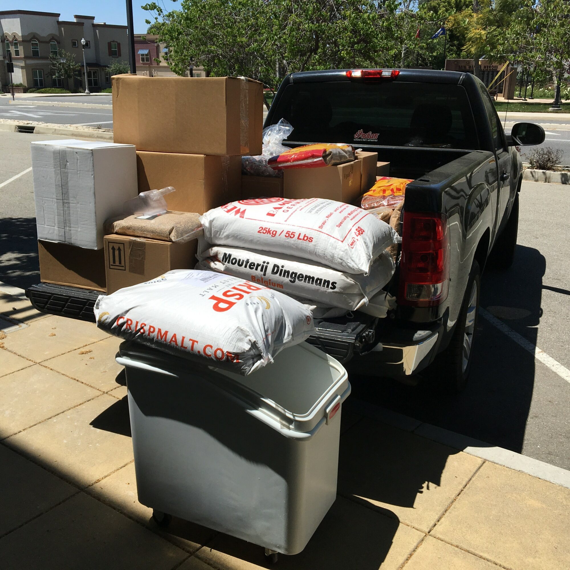 Pickup truck with boxes, bags, and a trash bin in the truck bed and on the ground, outdoors on a sunny day.