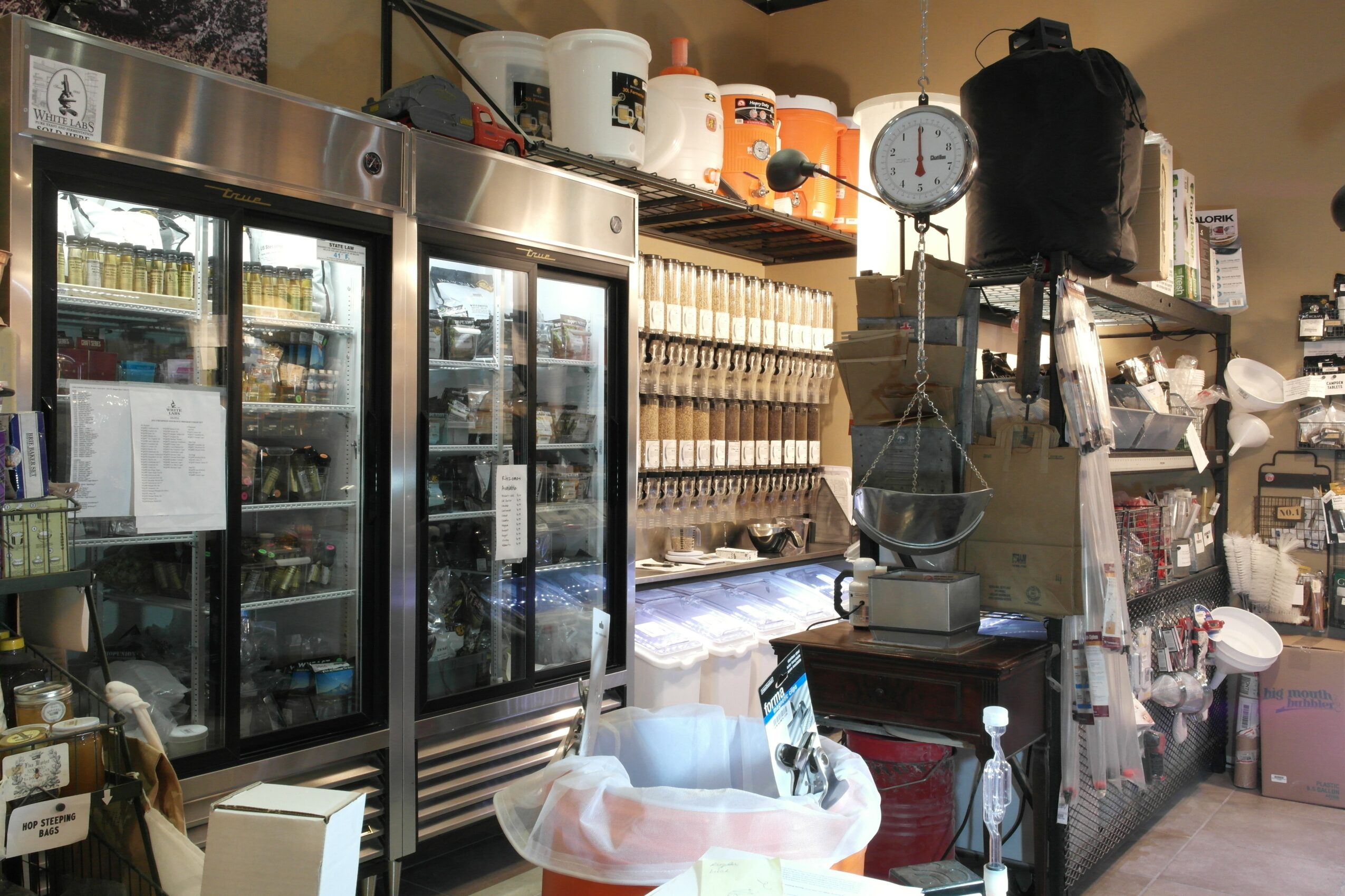 Commercial kitchen with refrigerators, shelves, and various kitchen equipment, including a scale and containers, in a well-organized space.