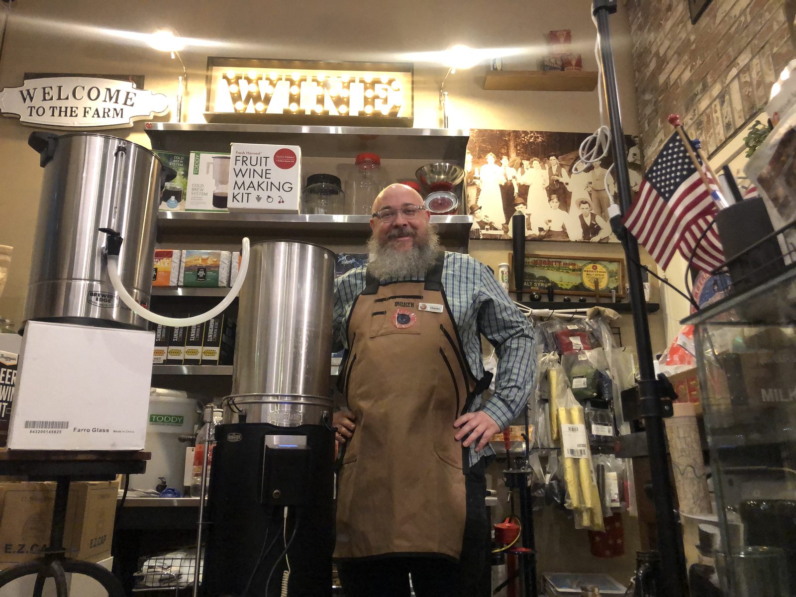 Smiling man with a beard wearing an apron behind a counter in a store, American flags and signs visible in background.