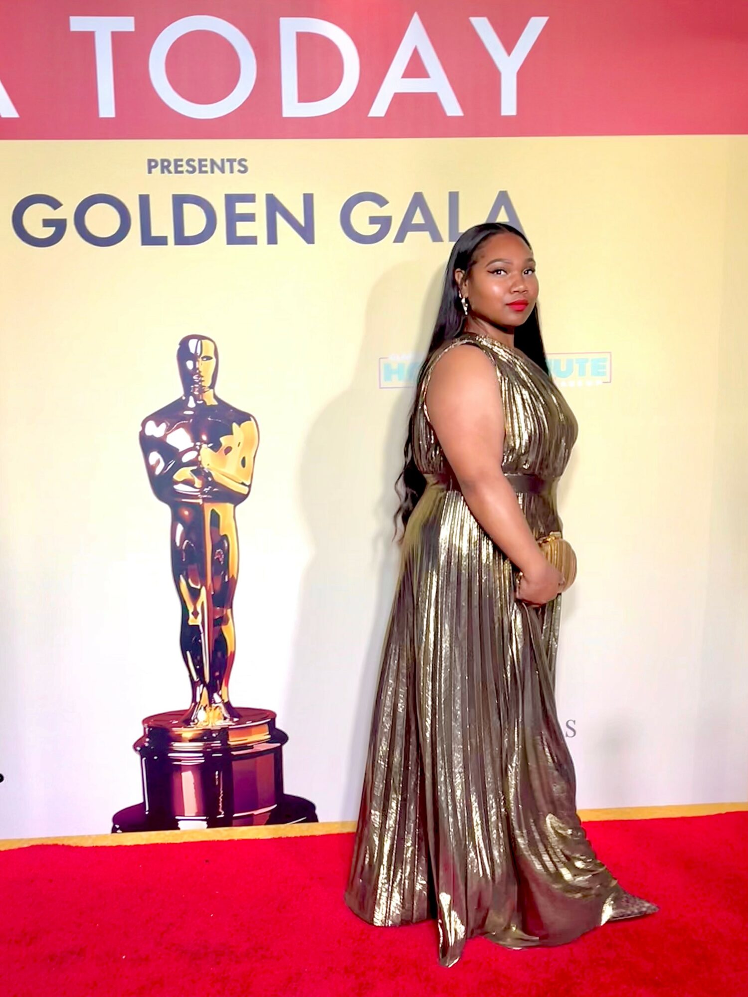 Woman in a metallic gold gown standing on red carpet at event, with a large Oscar statue and event signage in background.