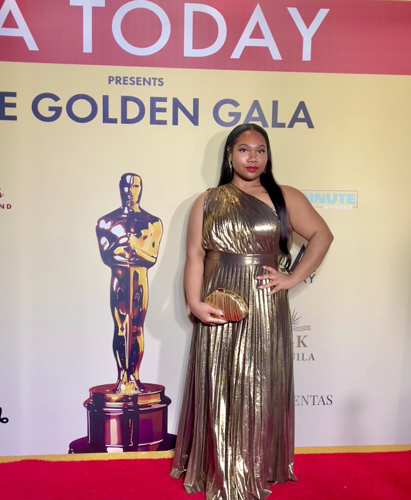 Woman in a gold metallic dress holding a clutch, standing on a red carpet in front of a yellow backdrop with an Oscar statue image.