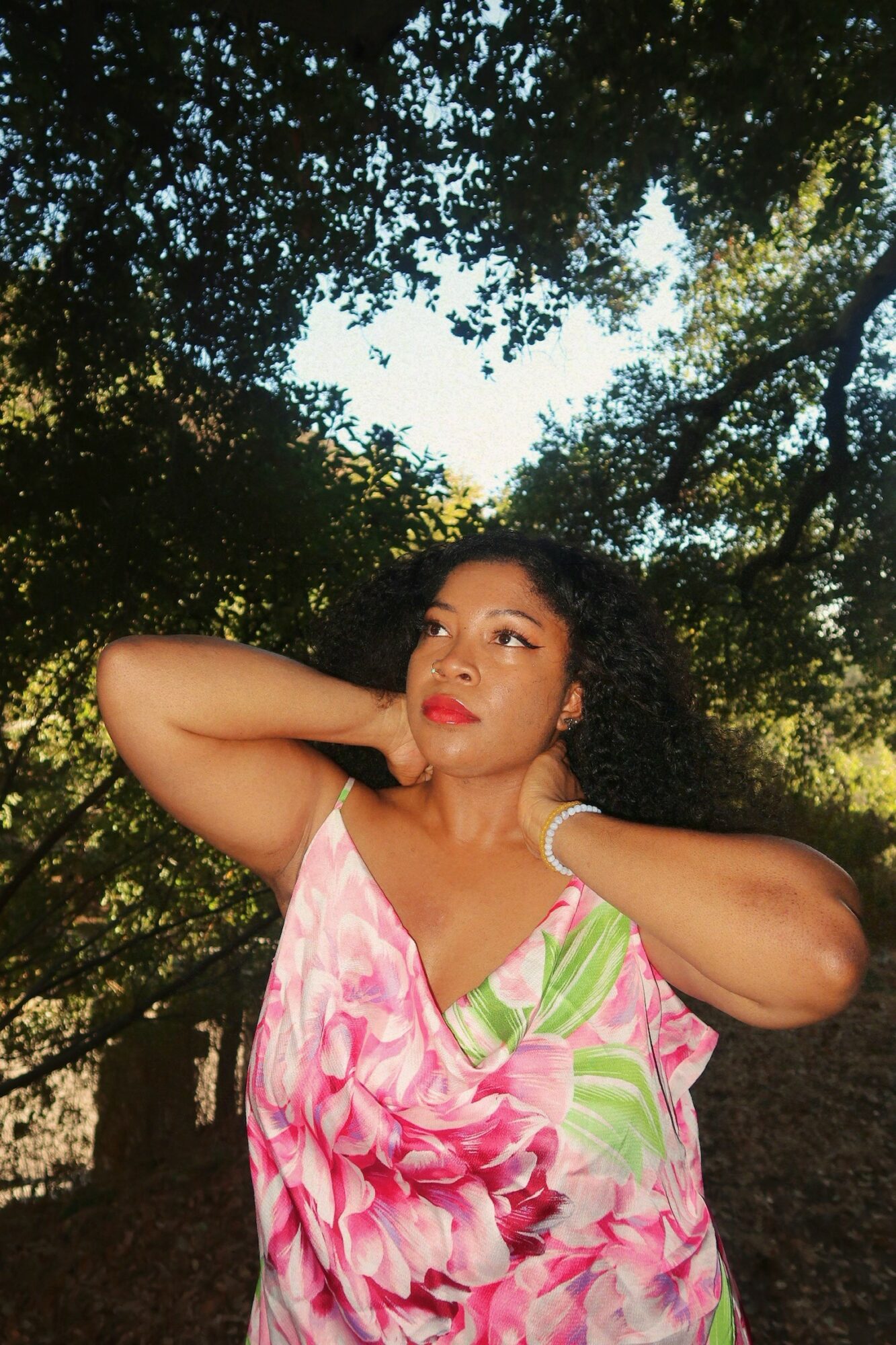 Woman with curly hair in a pink and green dress outdoors, looking to the side with trees overhead.