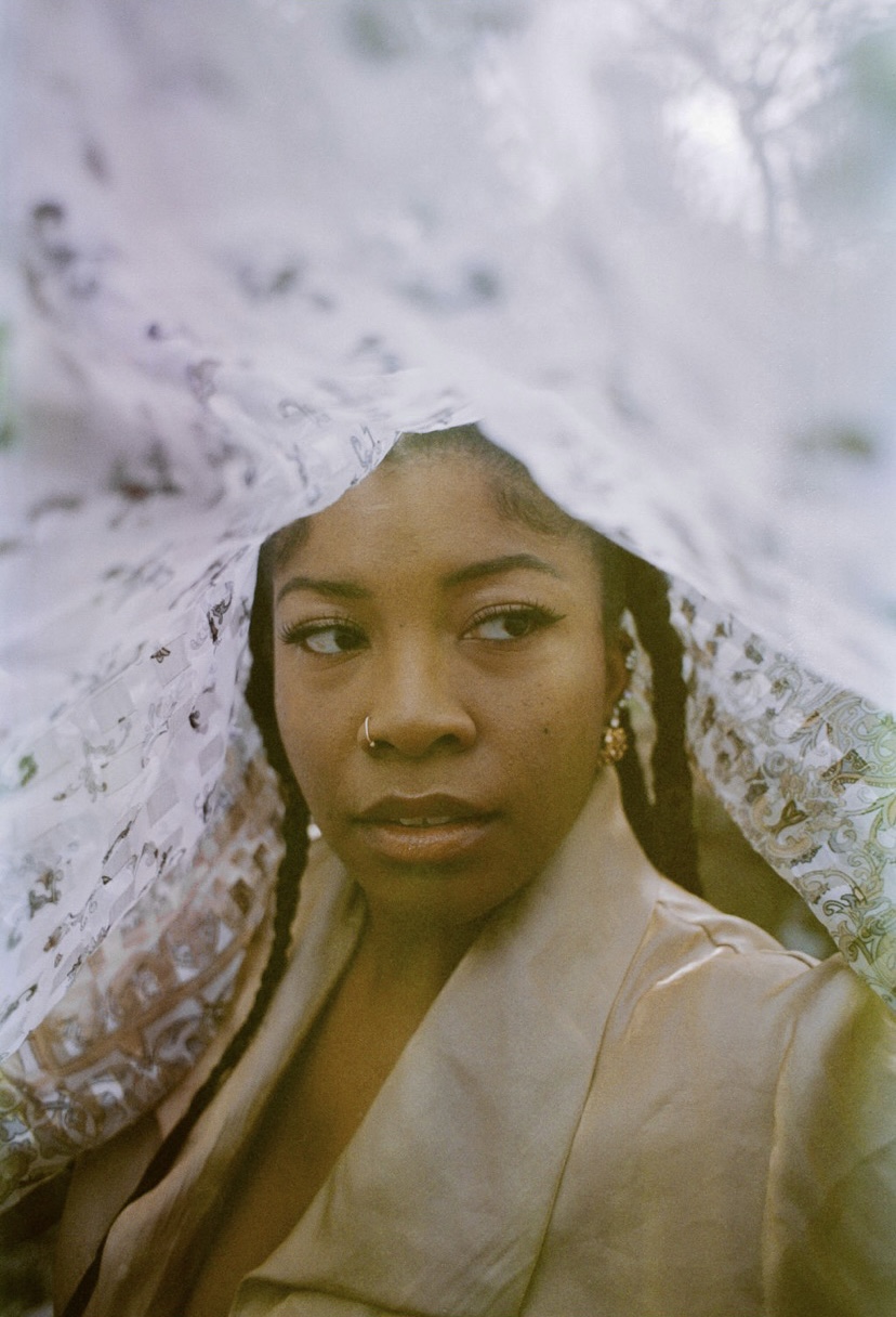 A woman with braided hair and earrings, wearing a beige jacket, looks to the side with a lace fabric above her head.