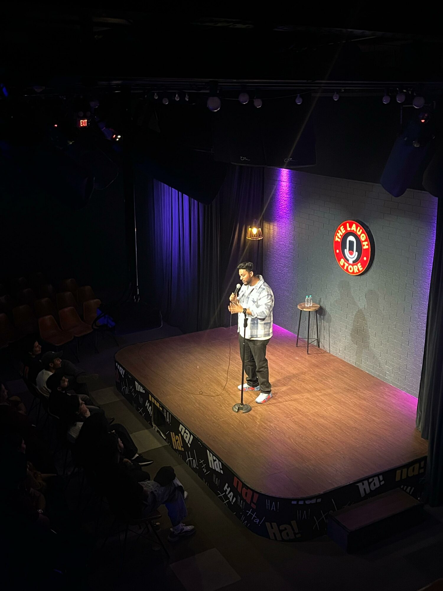 Person standing on stage with microphone, audience seated in front, stage lit with purple and yellow lights, comedy club logo on wall