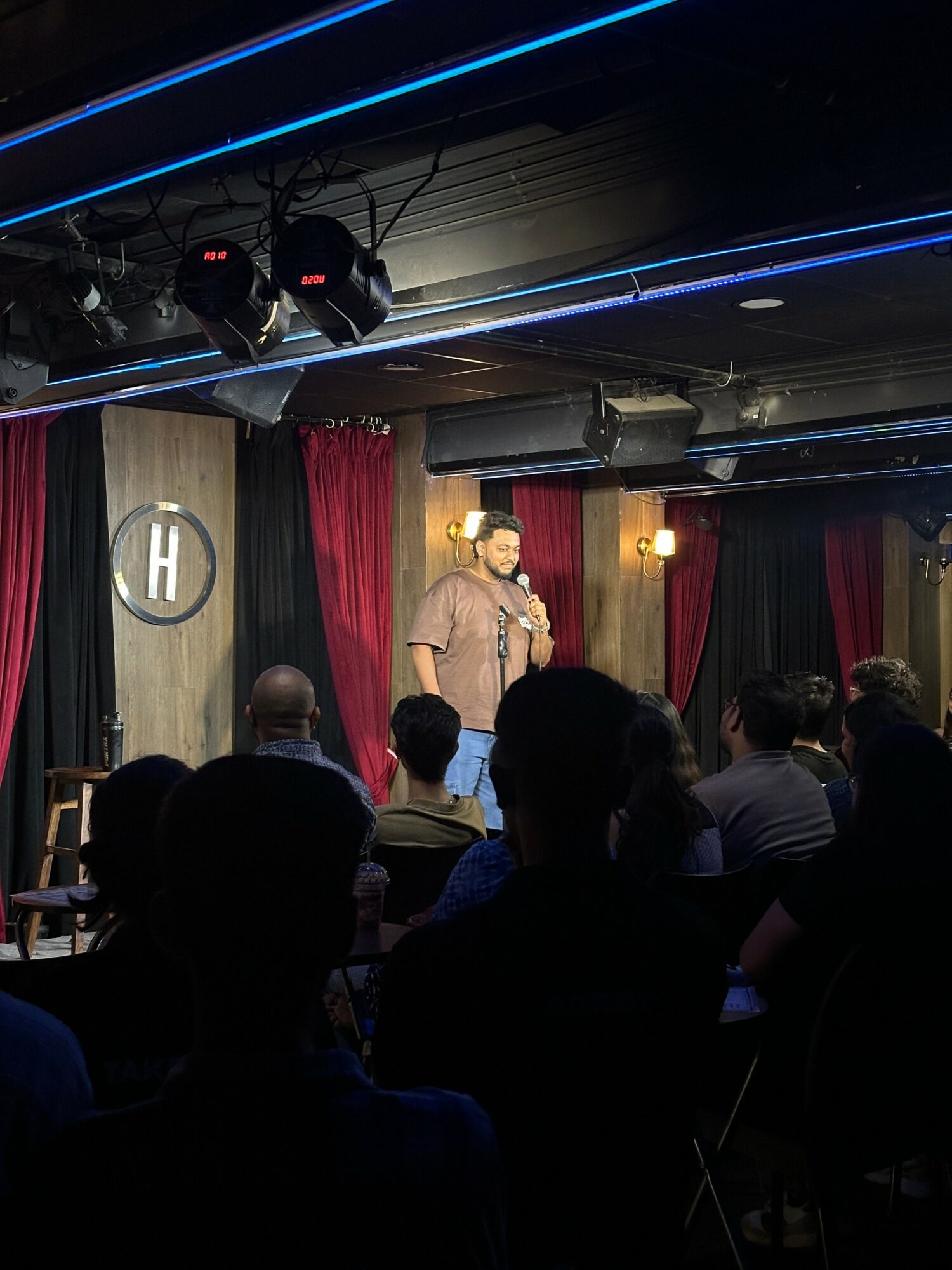 Stand-up comedian performs on stage with red curtains and audience watching, in a dimly lit comedy club.