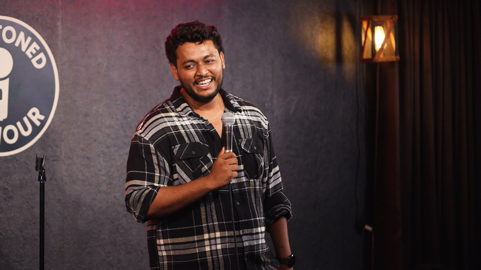 Person holding a microphone on stage, smiling, with a dark background and a lamp, in a comedy club setting.