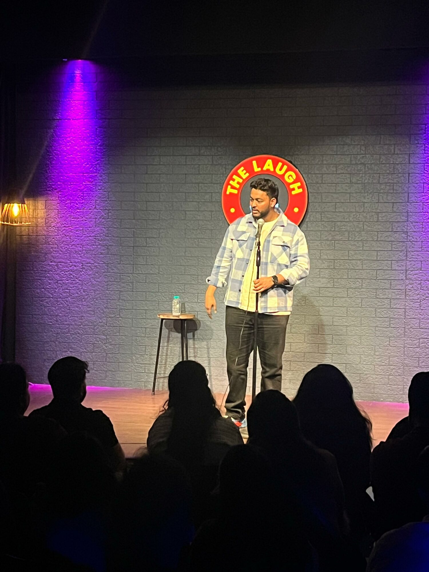 Stand-up comedian on stage with a microphone, a small table, and a glass of water, performing in front of an audience.