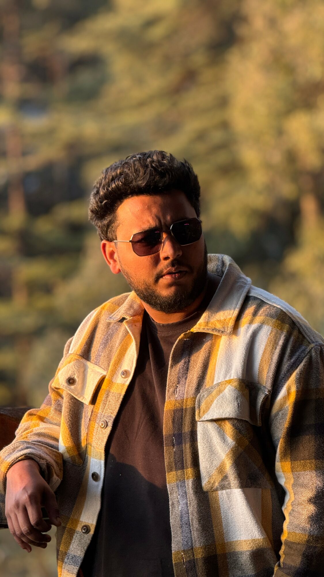 Man with sunglasses and curly hair wearing a plaid shirt outdoors with trees in the background.