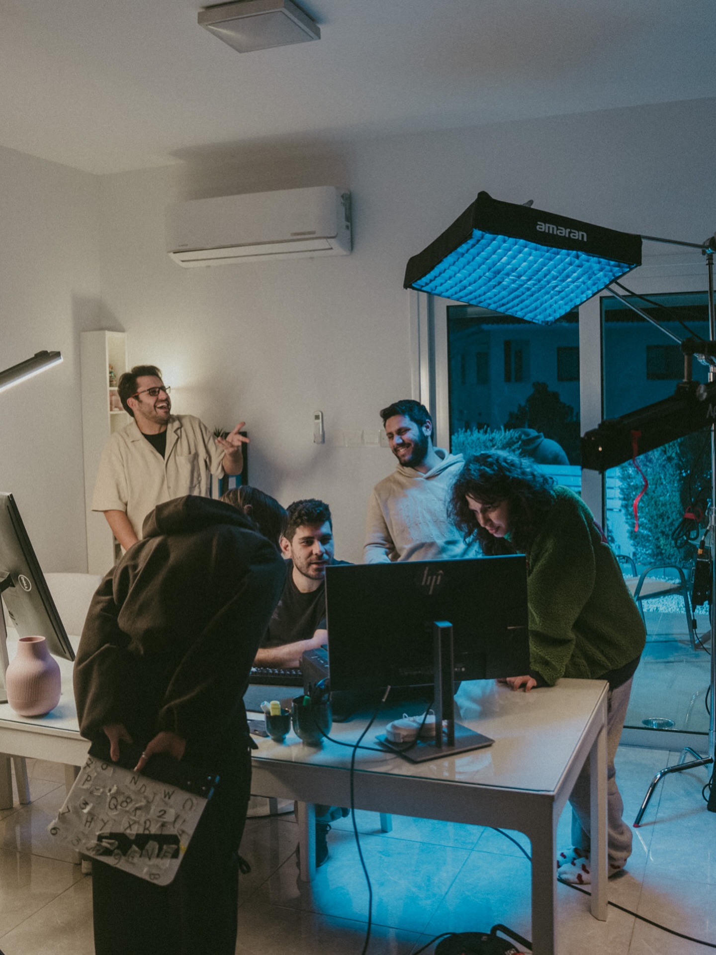 Group of five people gathered around a computer in a room with a large light, window, and air conditioning unit.