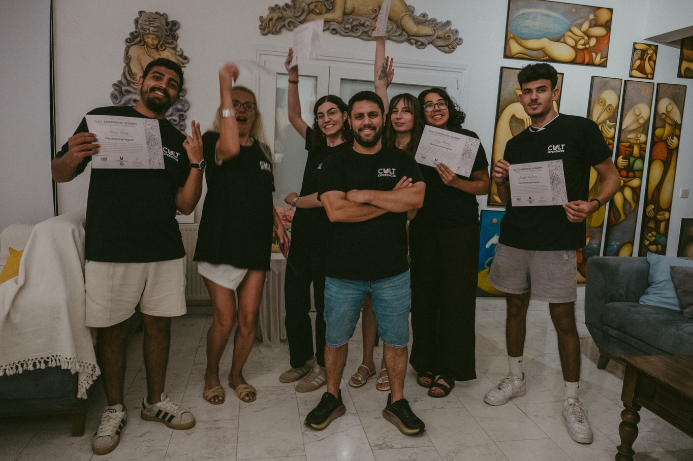 Group of six people celebrating, some holding certificates, smiling, in a room with colorful artwork on the walls.