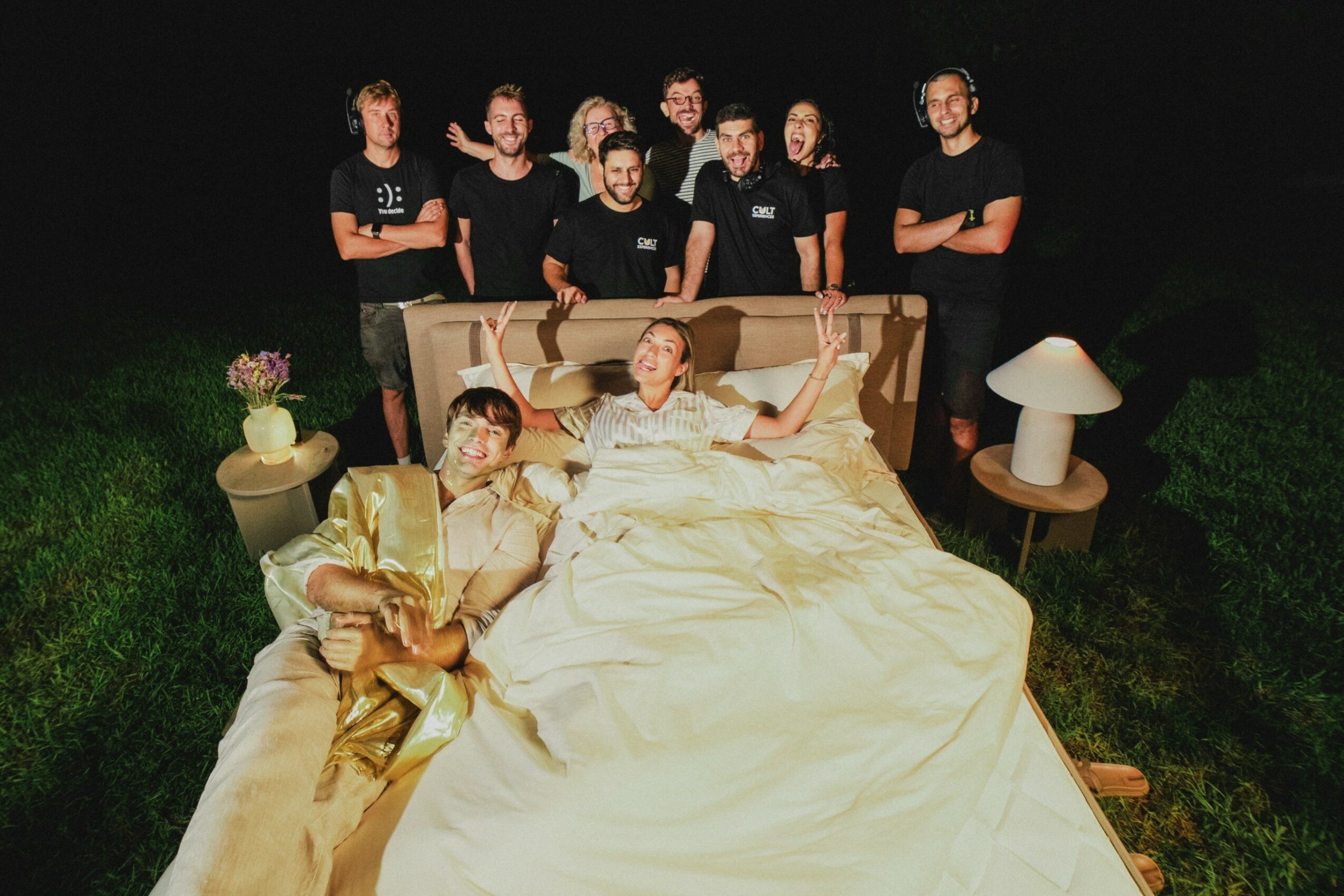Group of people standing behind a bed with two individuals lying on it, outdoors at night, with lamps on side tables.