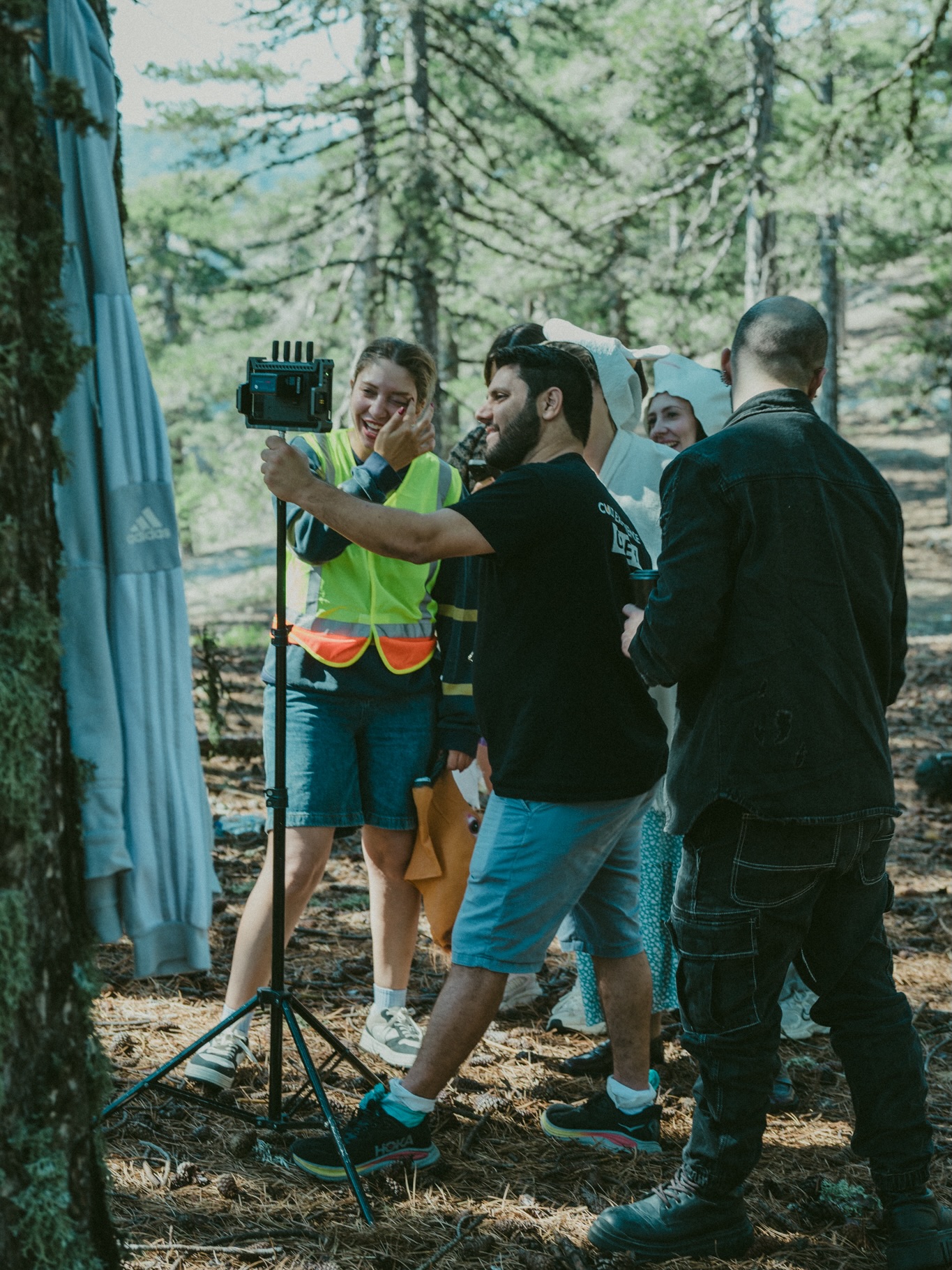 Group of people in a forest, one person holding a camera on a tripod, others watching, trees in background.