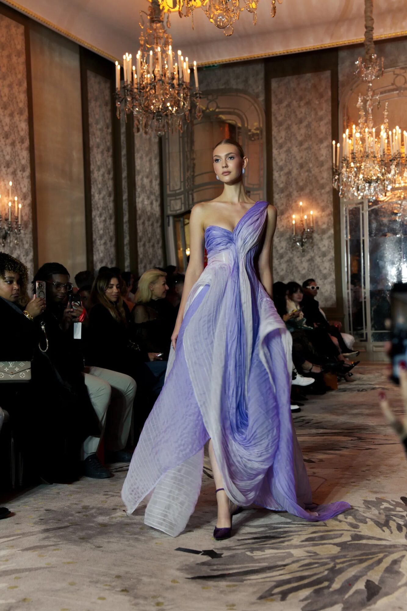 Model walking on runway in elegant purple and white gown with chandeliers and audience in background.