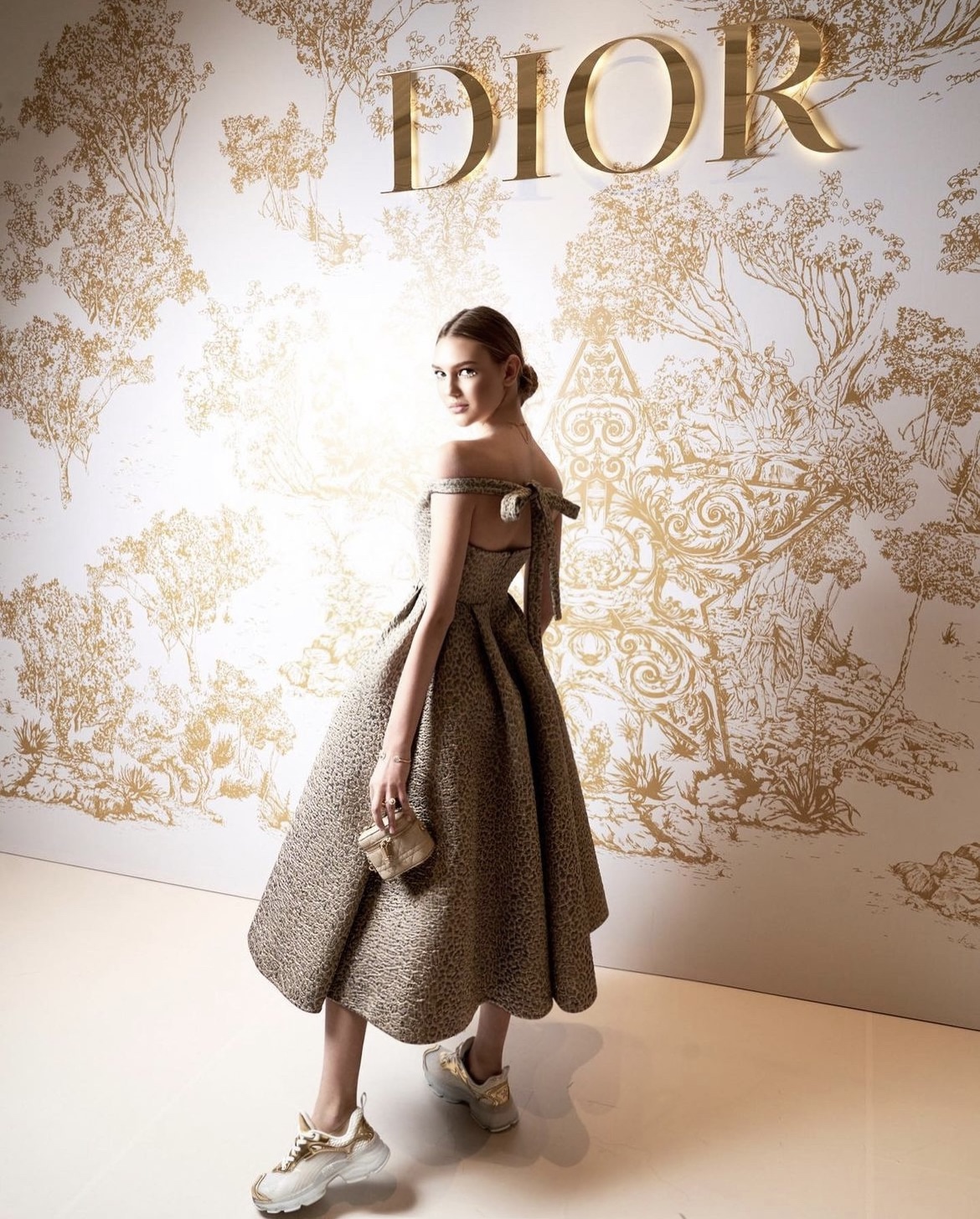 Woman in a dress holding a clutch standing in front of a Dior backdrop with gold illustrations.