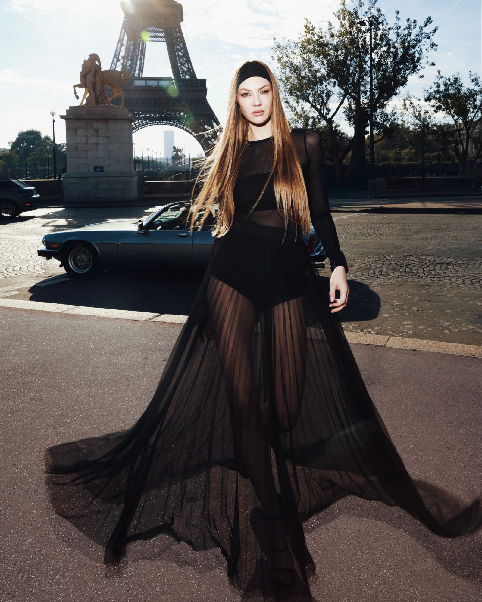 Young woman with long hair in black sheer dress standing on street with Eiffel Tower replica in background.