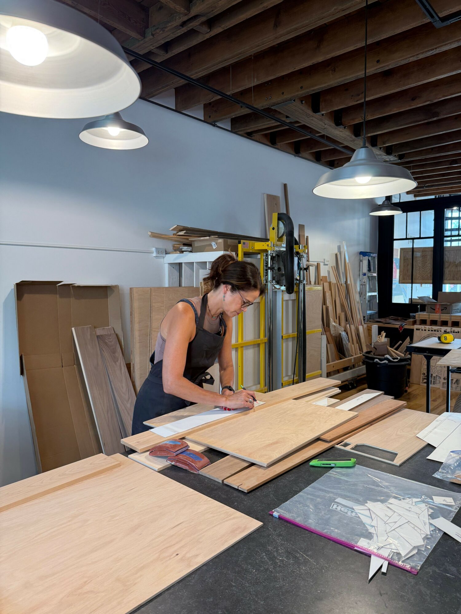 Woman working at a woodworking table in a workshop with tools and materials, natural light from windows, ceiling lights overhead.