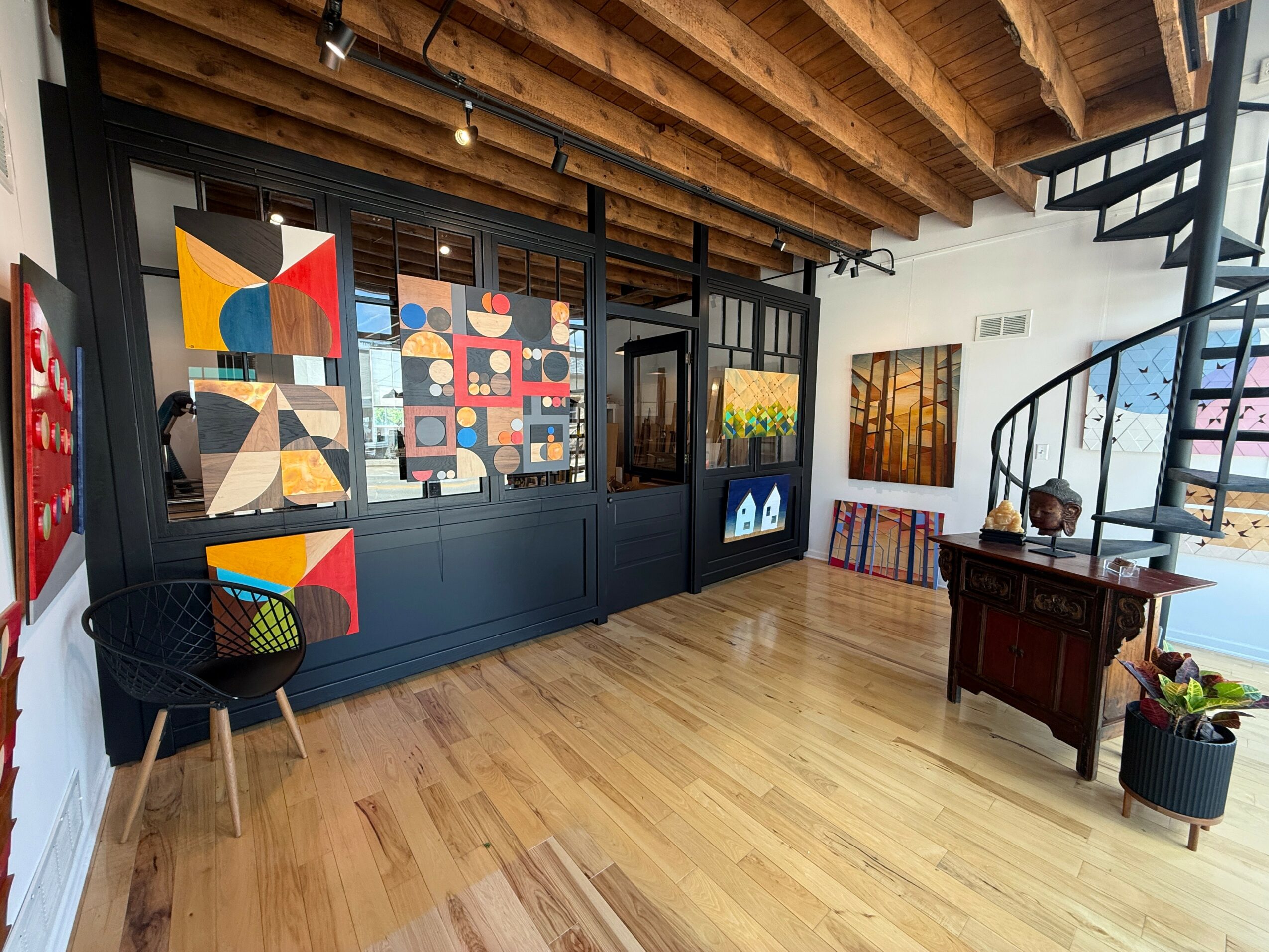 Art gallery interior with colorful paintings on dark wall, wooden ceiling, spiral staircase, and wooden floor.
