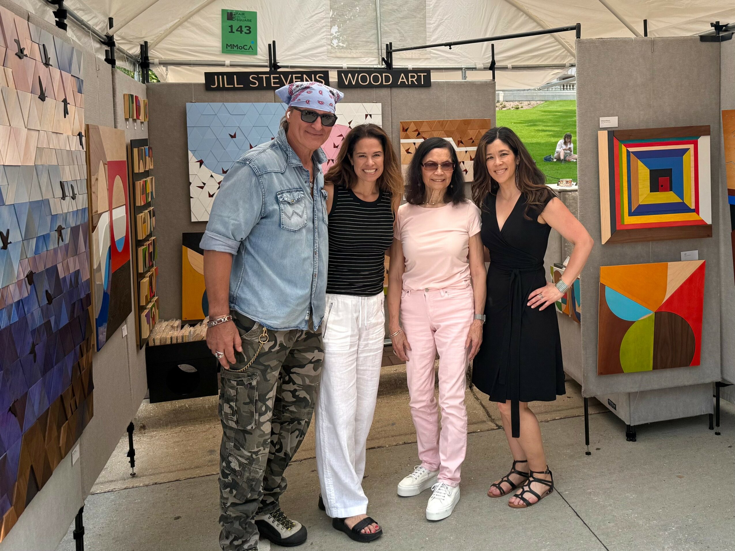 Four people stand together at an art booth, smiling, with colorful artwork displayed behind them.