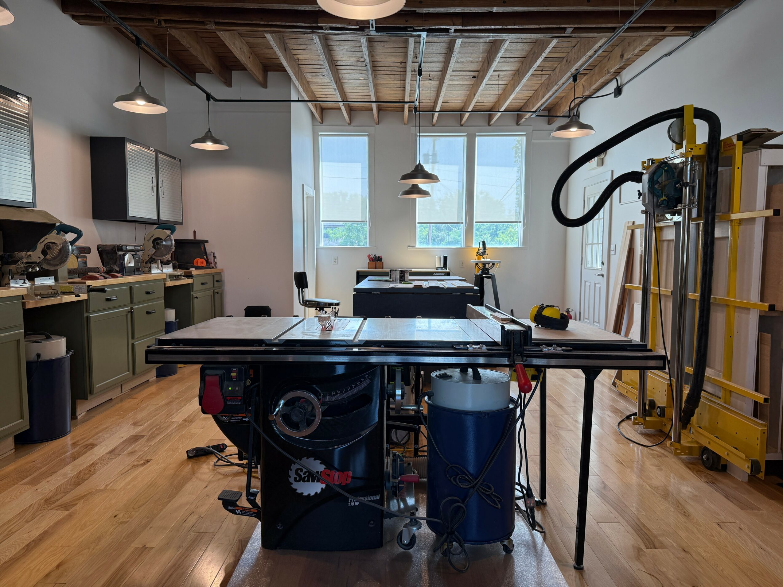Table tennis table in a room with large windows, kitchen area, and exercise equipment, wooden floor, and ceiling lights.