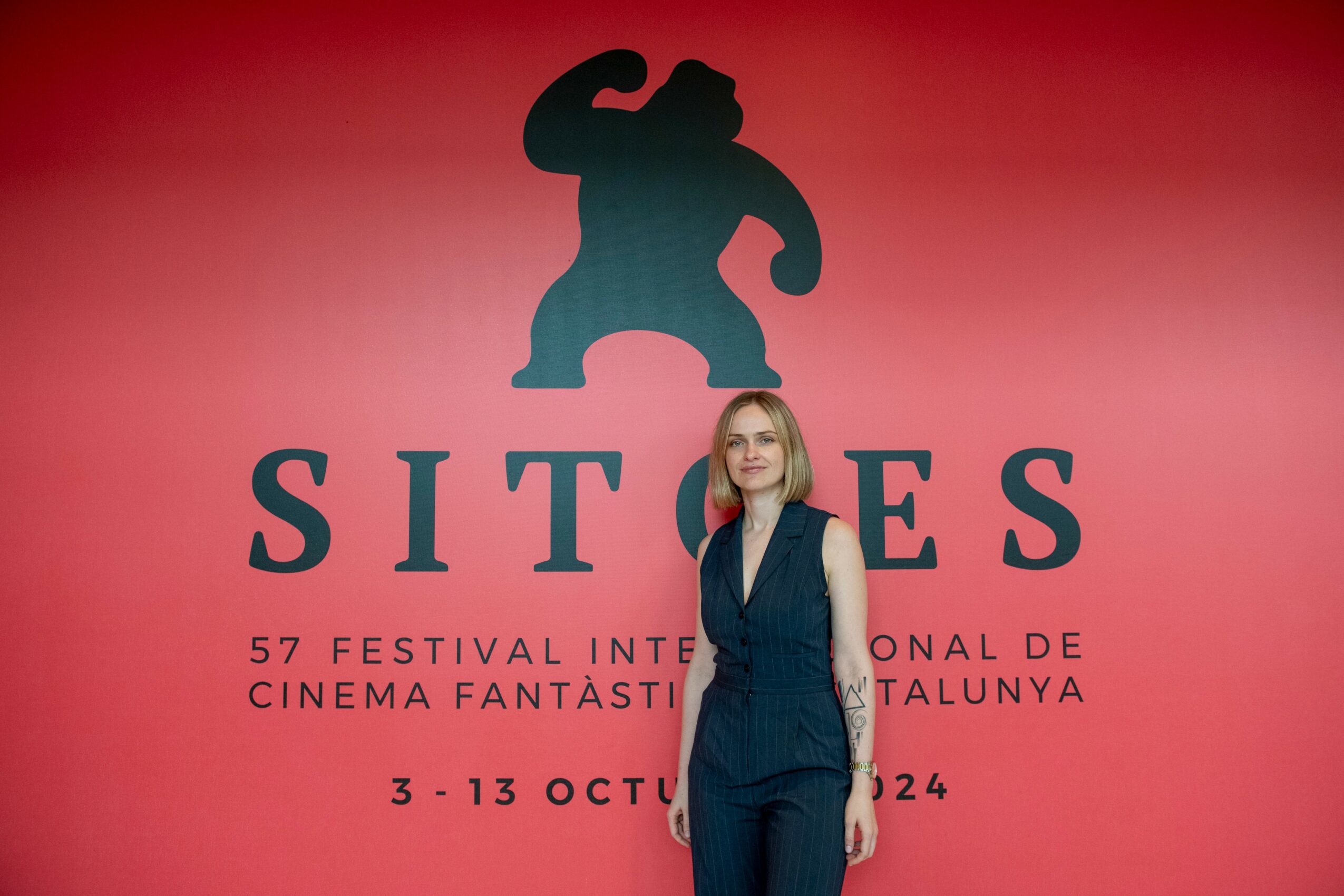 Woman with blonde hair in a dark blue vest and pants standing in front of a red background with a bear silhouette and text.