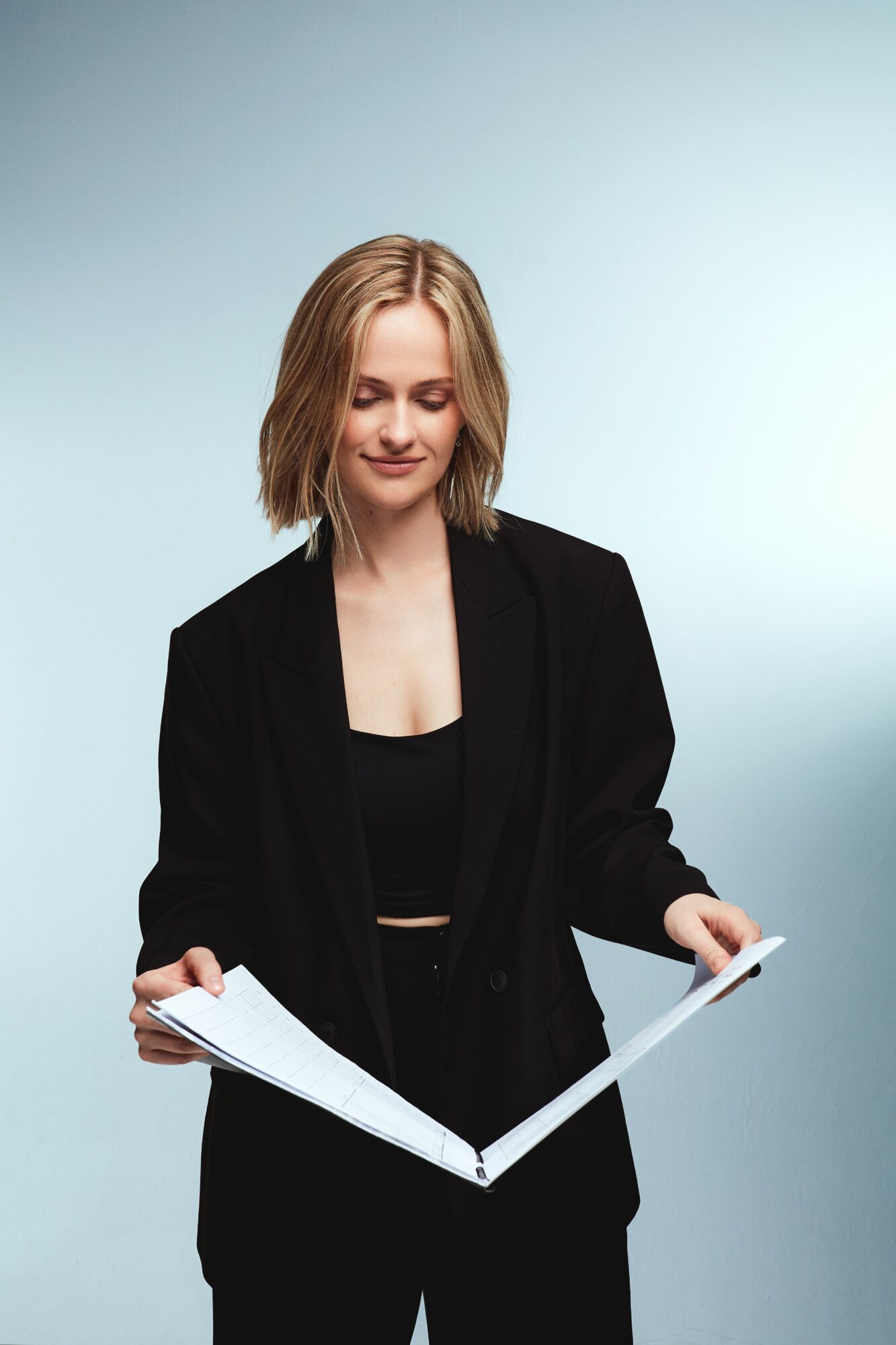 Young woman with shoulder-length blonde hair wearing a black blazer and top, holding an open book, smiling softly.
