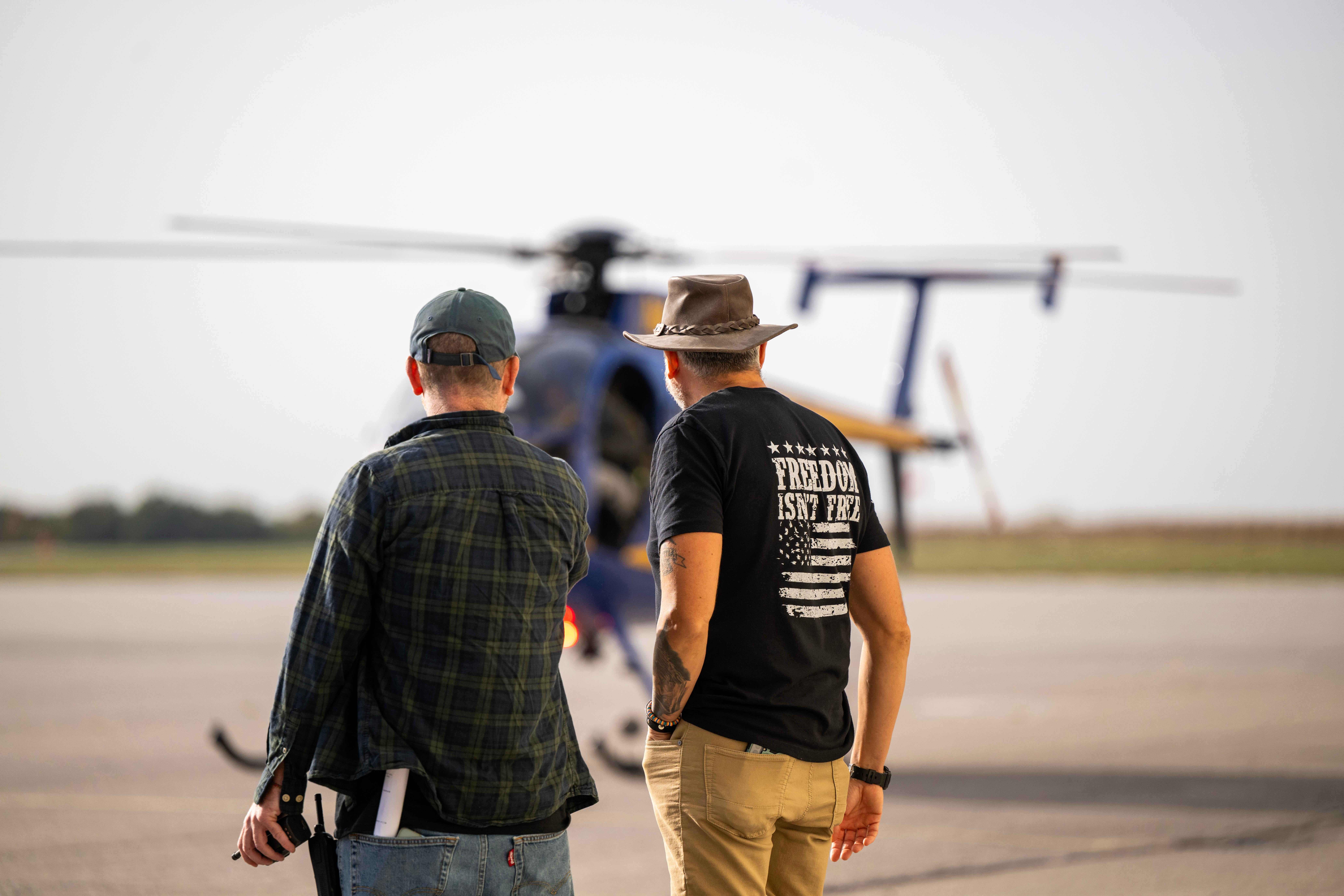 Two men stand on tarmac watching a helicopter take off or land, with a clear sky in the background.