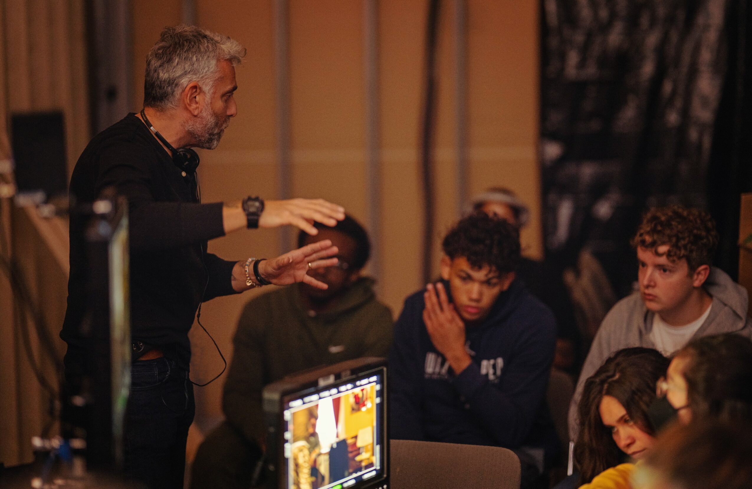 A man with gray hair and beard gestures while speaking to a group of young people seated around a table.