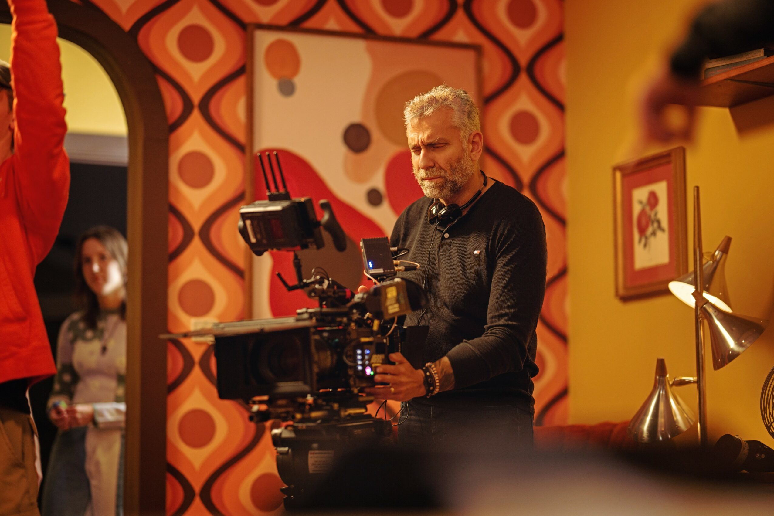 Man operating a camera in a colorful room with patterned wallpaper and framed artwork, with a woman in the background.