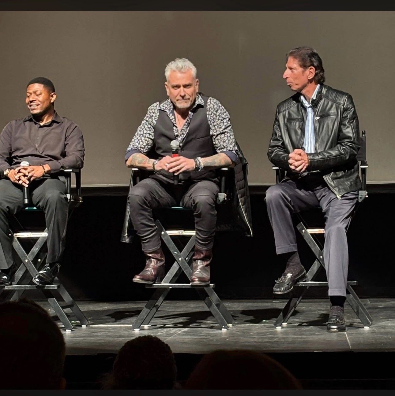Three men sitting on chairs on stage, engaged in conversation, with a plain background and audience visible at the bottom.