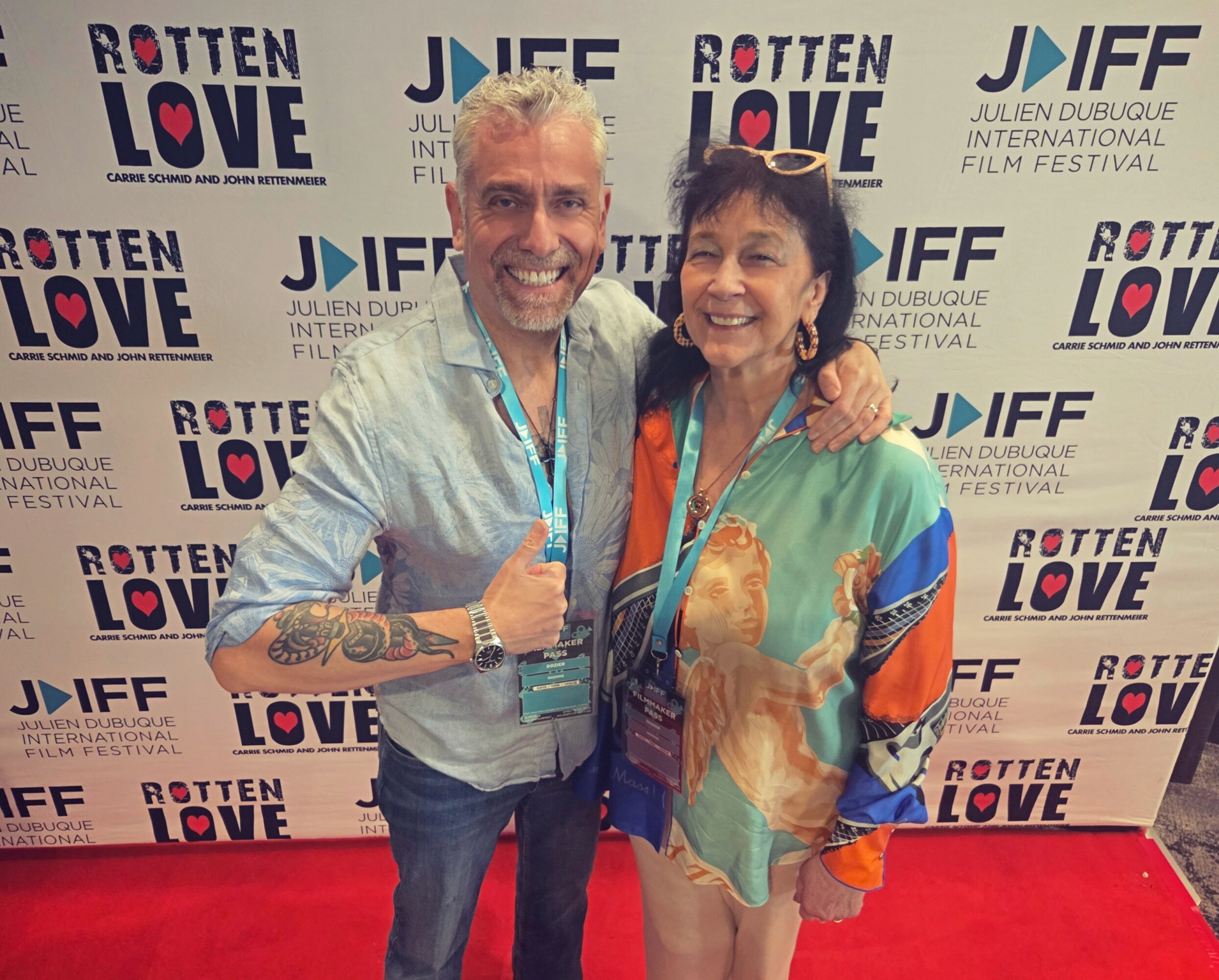 Two smiling people with lanyards pose in front of a backdrop with event logos, one with arm around the other.