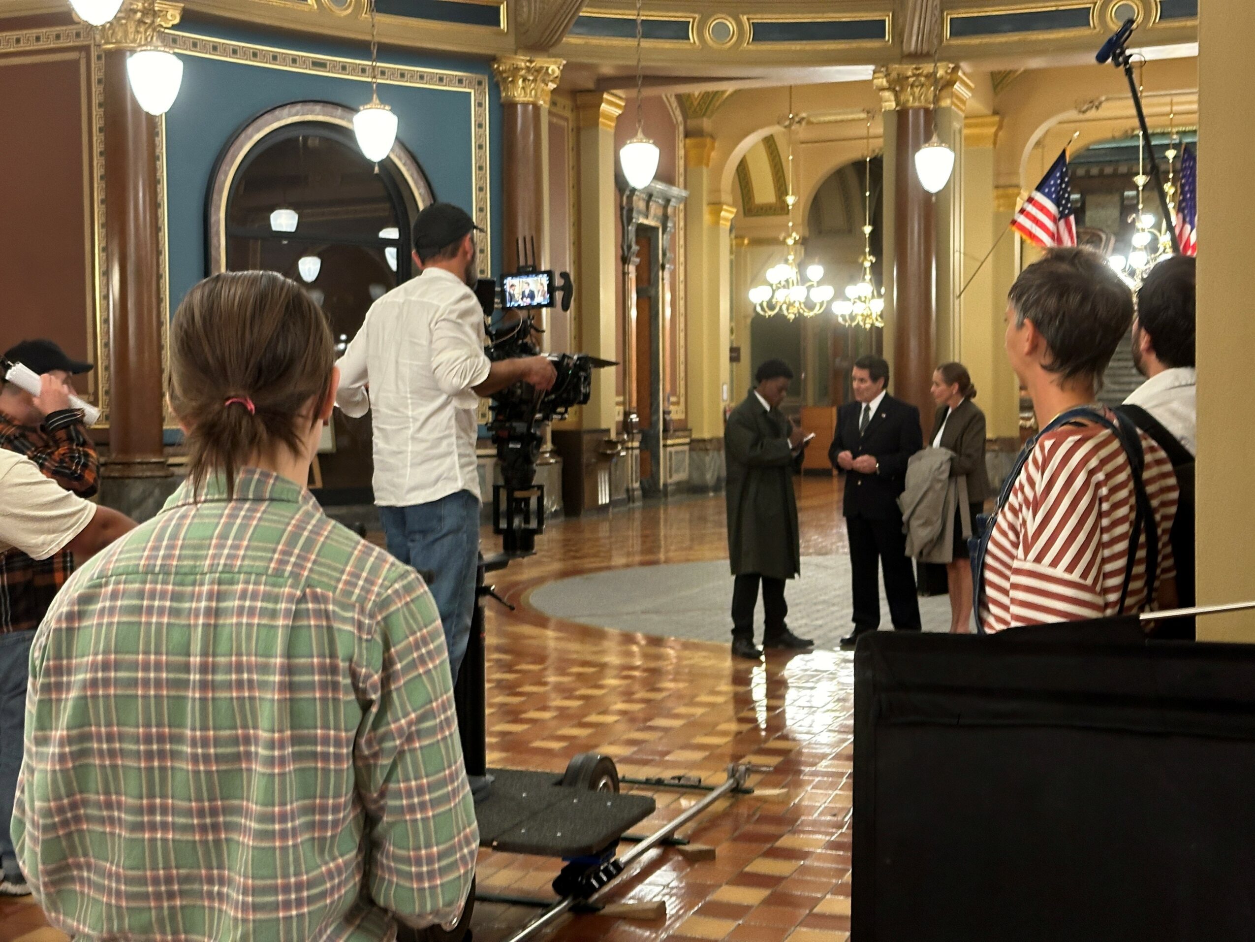People filming and observing a formal event in an ornate, well-lit room with gold accents and American flags.