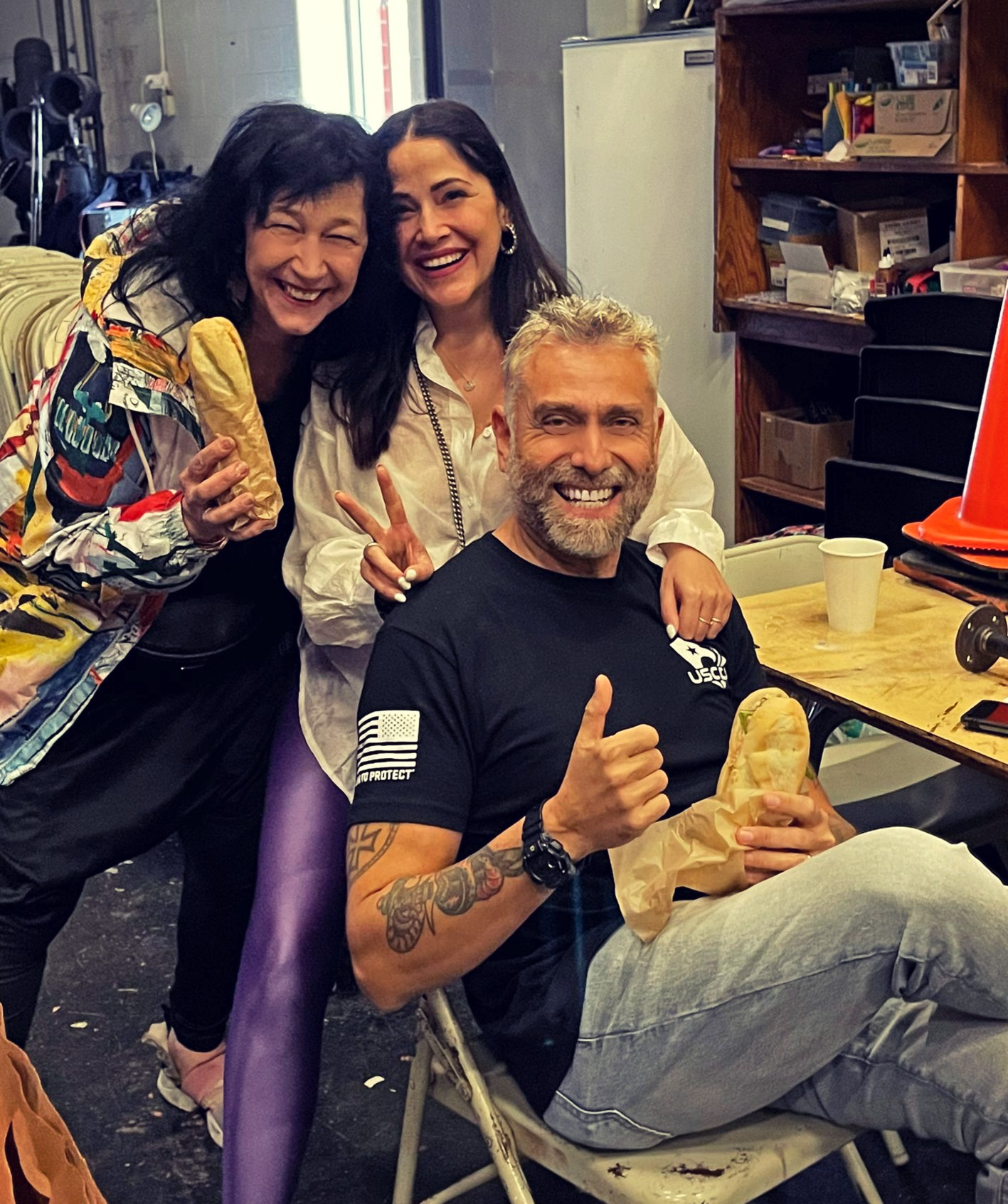 Three people smiling and posing together indoors, one seated and two standing behind, with shelves and a table in the background.