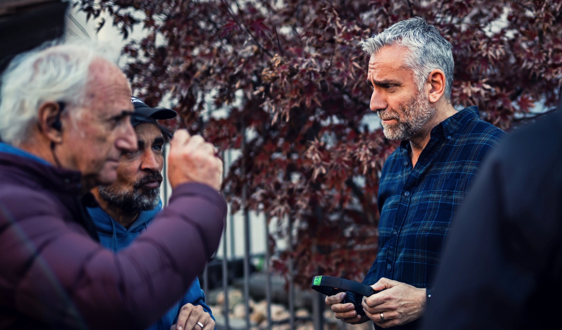 Group of people outdoors, engaged in conversation, with trees and a fence in the background.