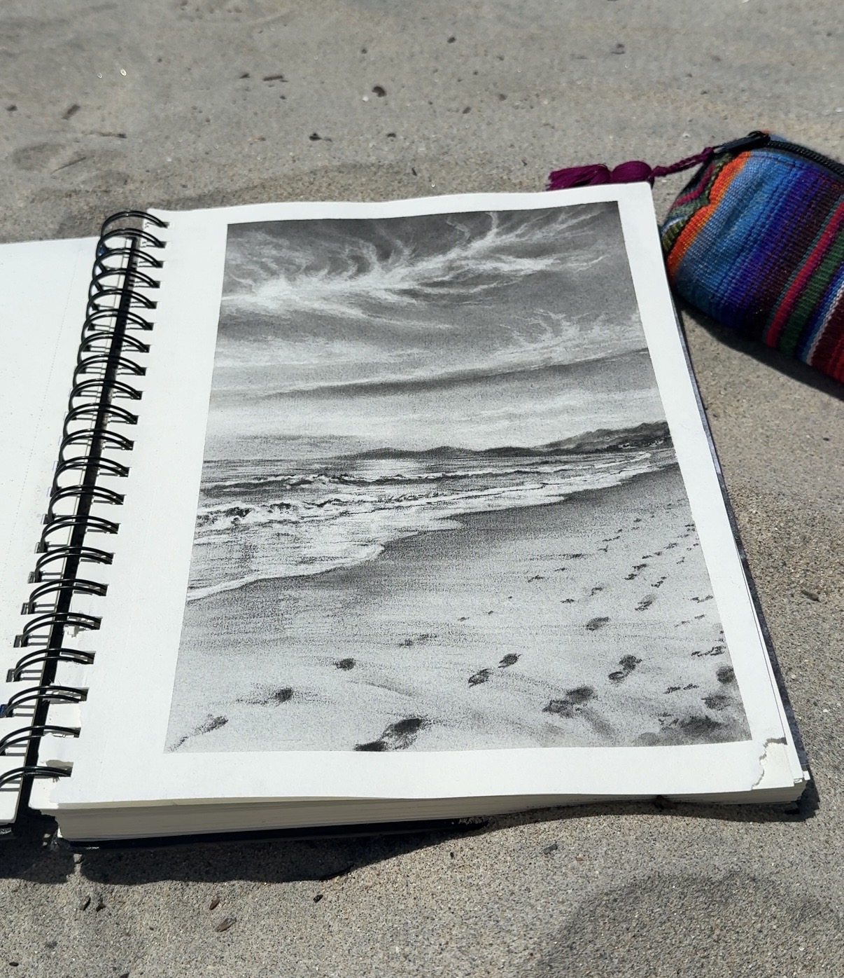 Black and white photo of a beach with waves and footprints, in a spiral notebook on sand, with a colorful bag nearby.