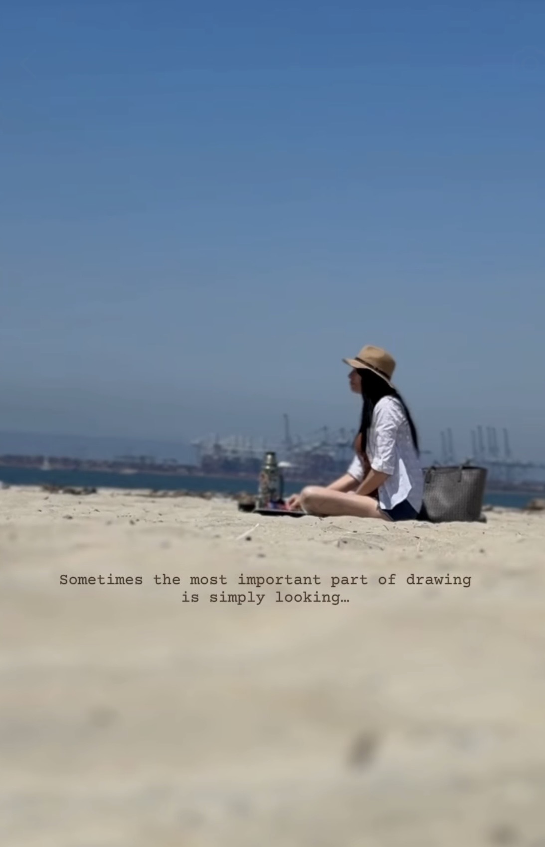 Person sitting on sandy beach with a hat, looking at the water and sky, with a cityscape in the distance.