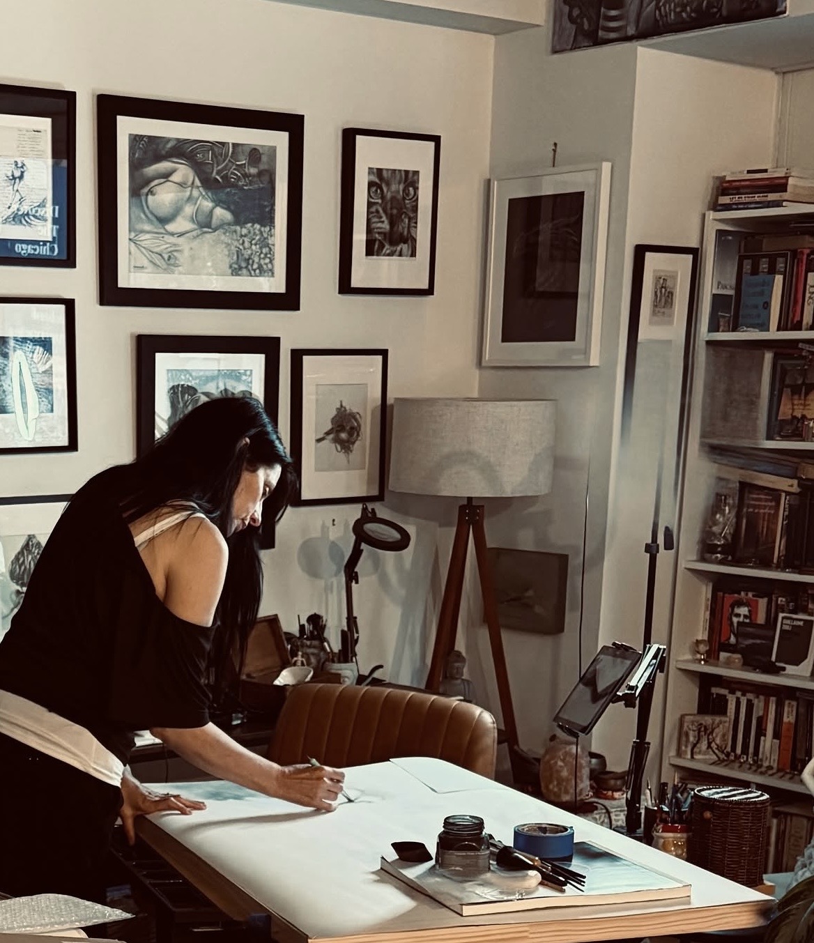 Woman writing on large paper in a room with framed pictures, bookshelf, and lamp.