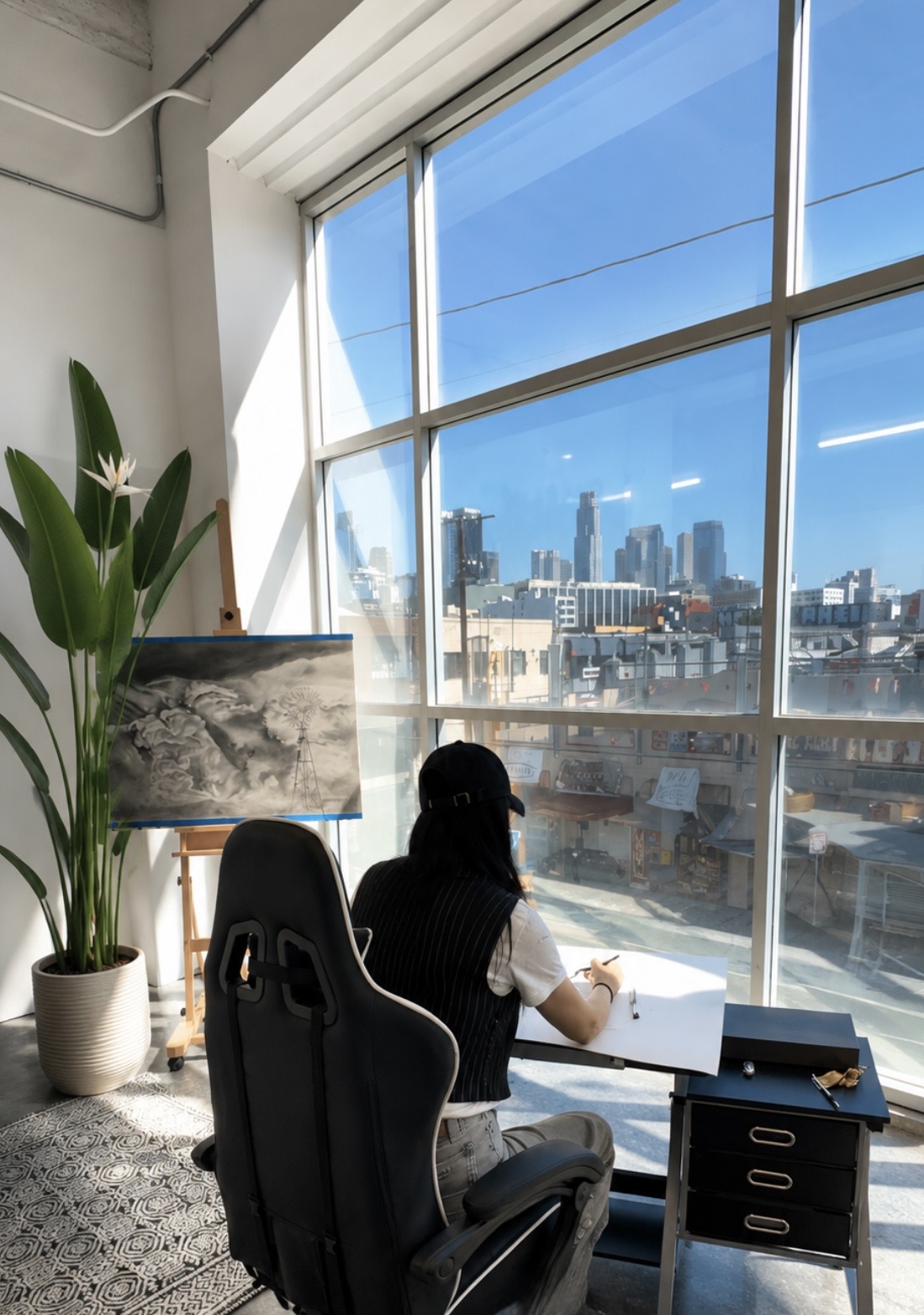 Person sitting at a desk by a large window overlooking a cityscape, with a potted plant nearby.