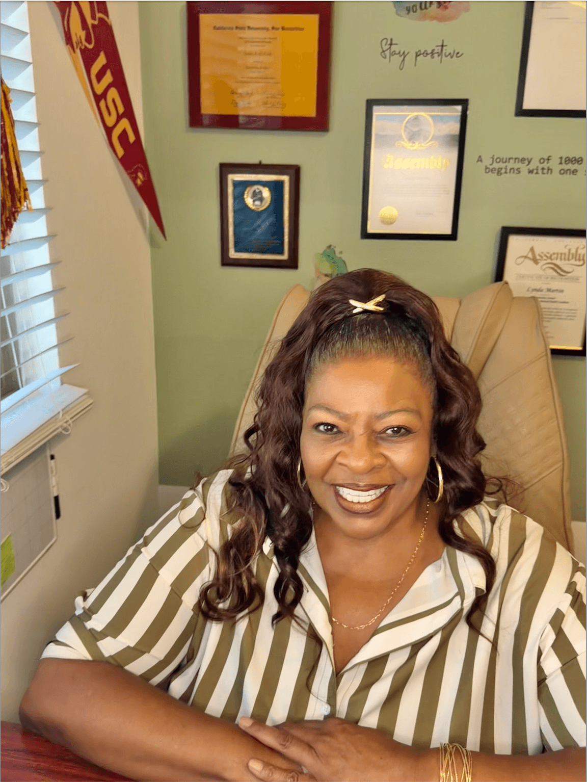 Smiling woman with curly hair sitting in an office chair, wearing a striped shirt, in front of a wall with framed certificates and awards.