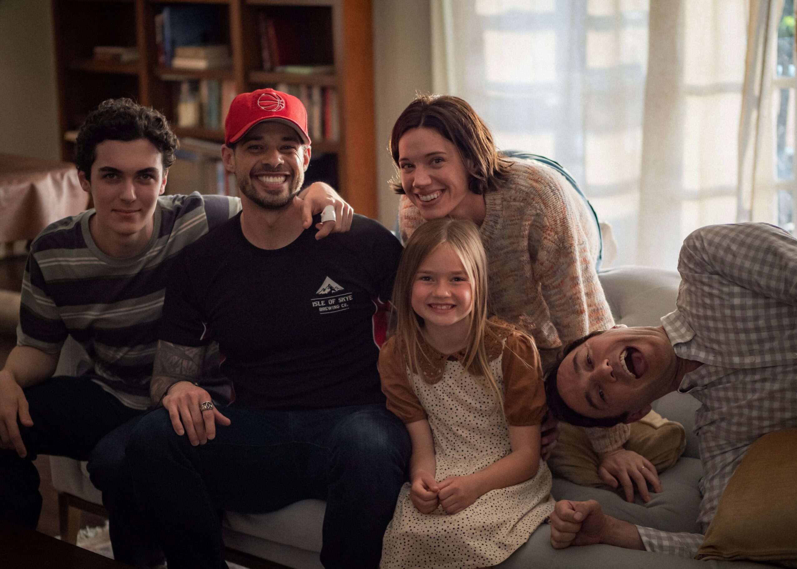 Group of five people sitting on a sofa, smiling indoors with sunlight through windows, including two children and three adults.
