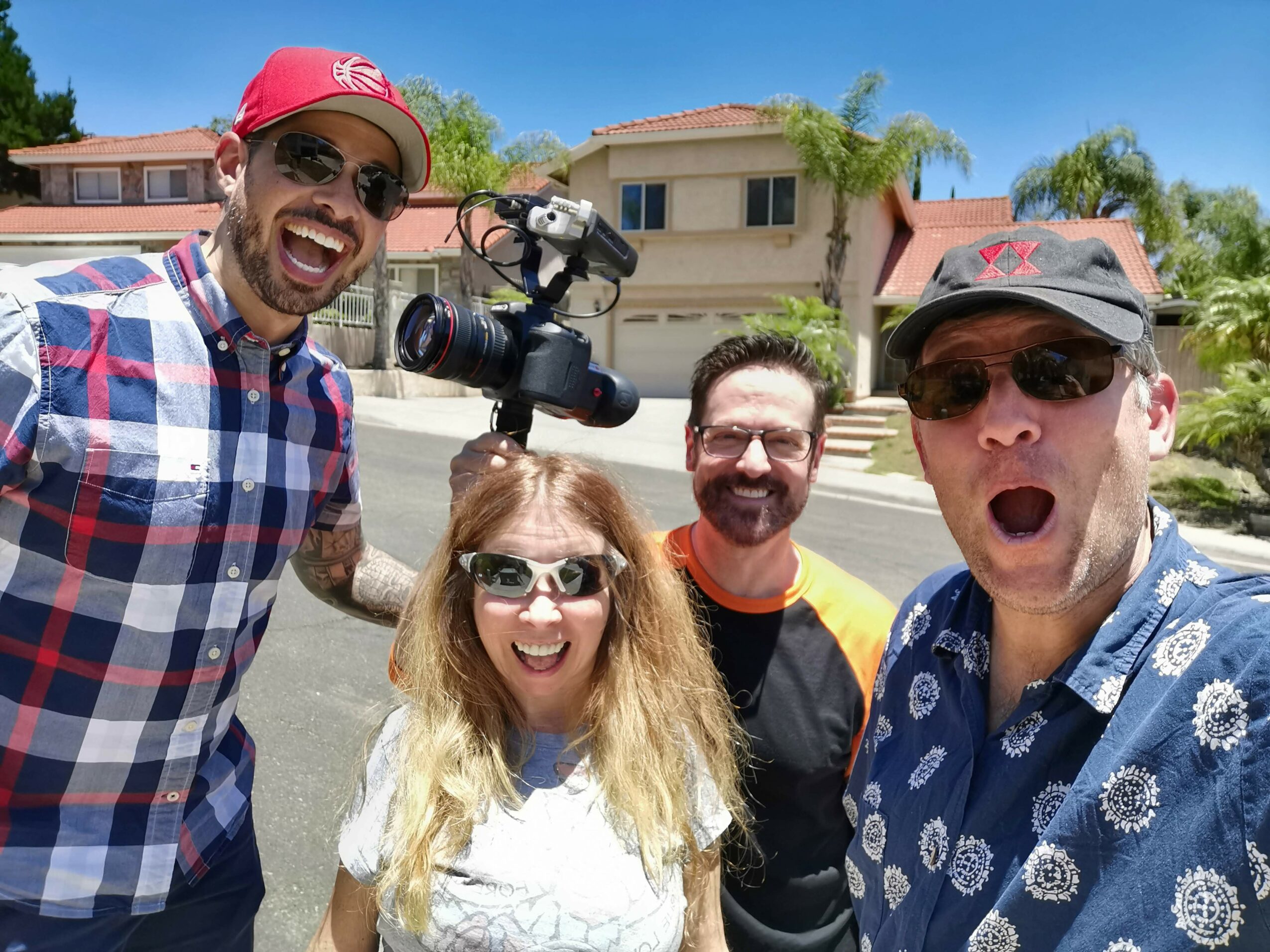 Four people smiling outdoors on a sunny day, with houses and trees in the background.