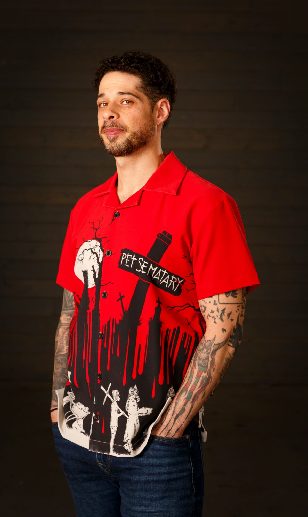 Young man with tattoos wearing a red shirt with graphic design, standing against a dark background.
