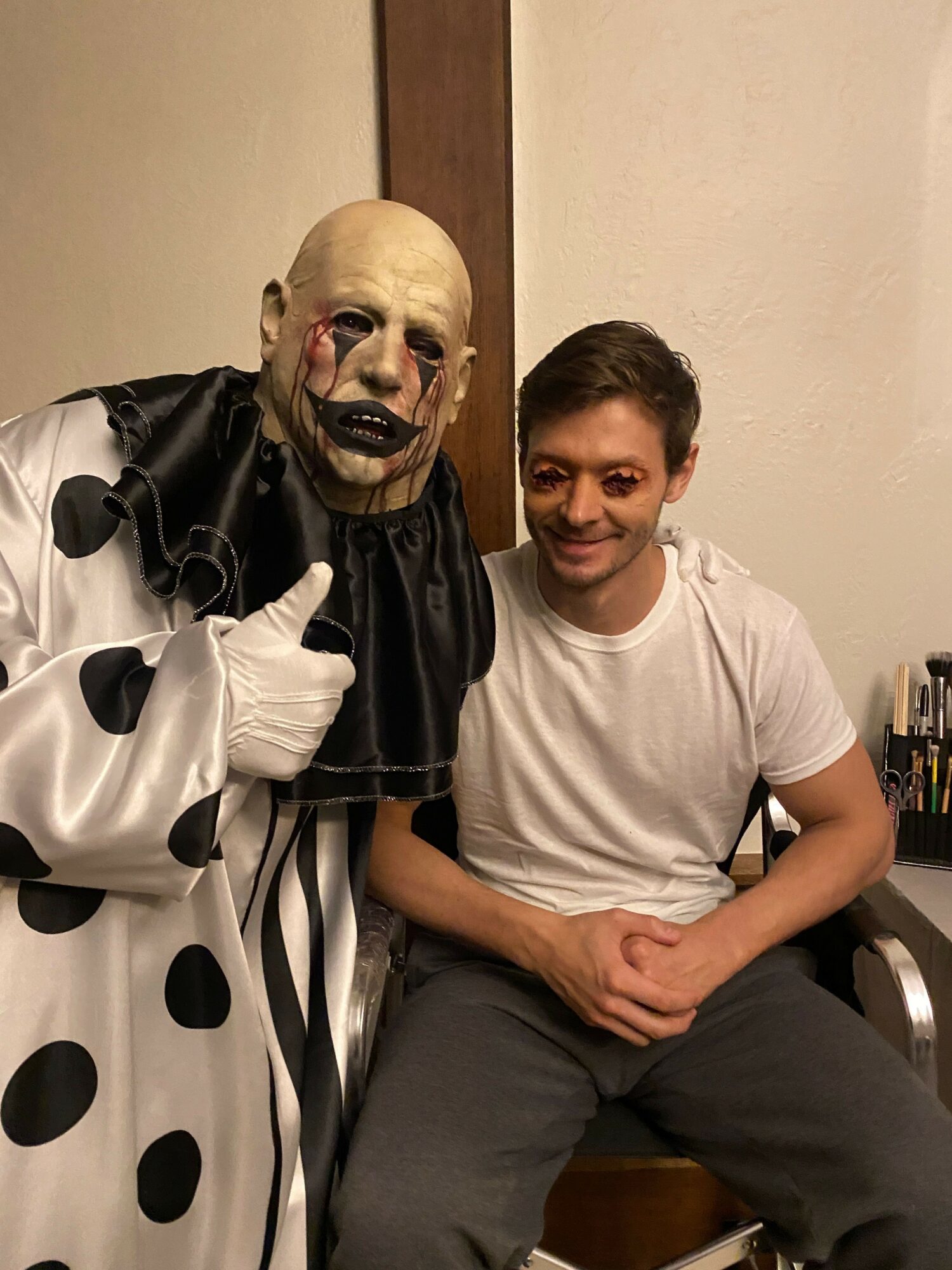 Person in a white t-shirt sitting next to a person in a clown mask, both seated indoors.