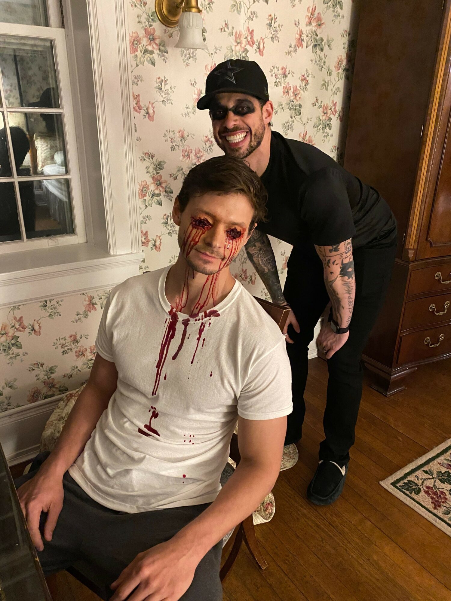 Person with blood on face and white shirt sitting at table, another person standing behind with tattoos, room with floral wallpaper
