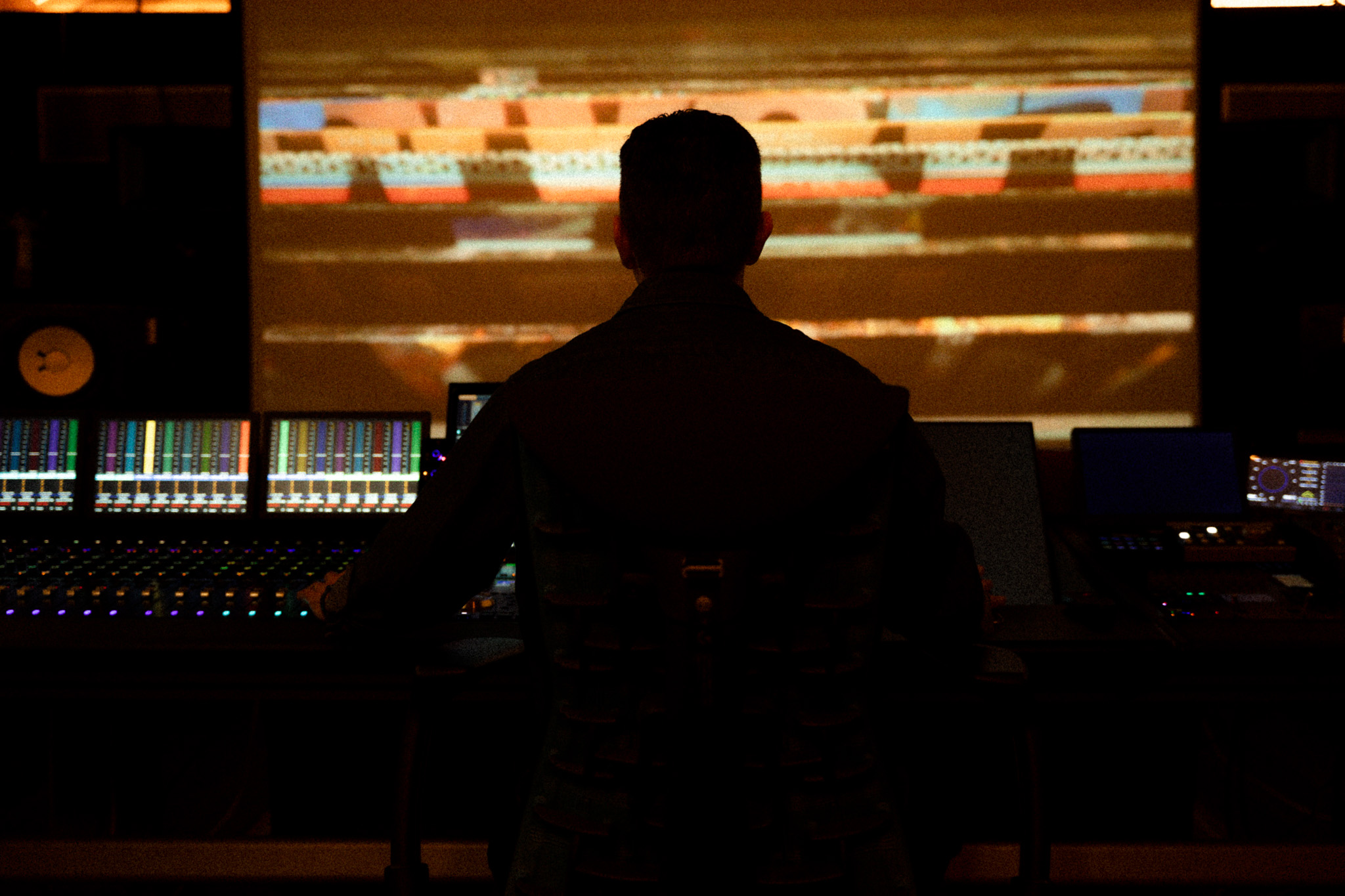 Person working at a mixing console in a dark room with a large screen displaying colorful visuals behind them.