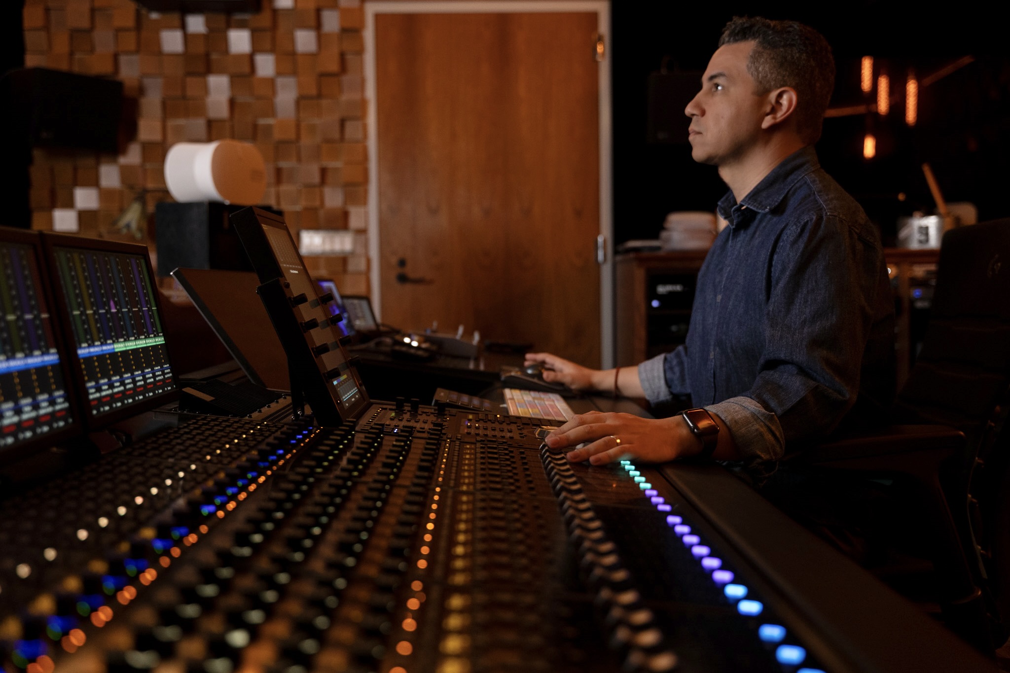 Man working at a sound mixing console in a recording studio, with equipment and wooden wall panels visible.