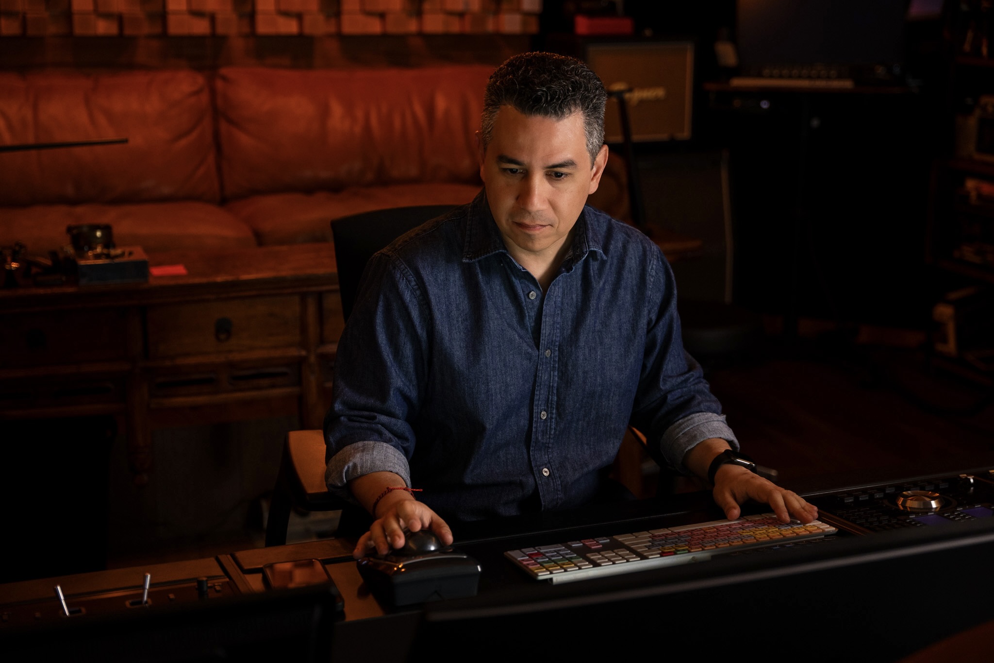 Man in dark blue shirt sitting at a control panel in a dimly lit room.